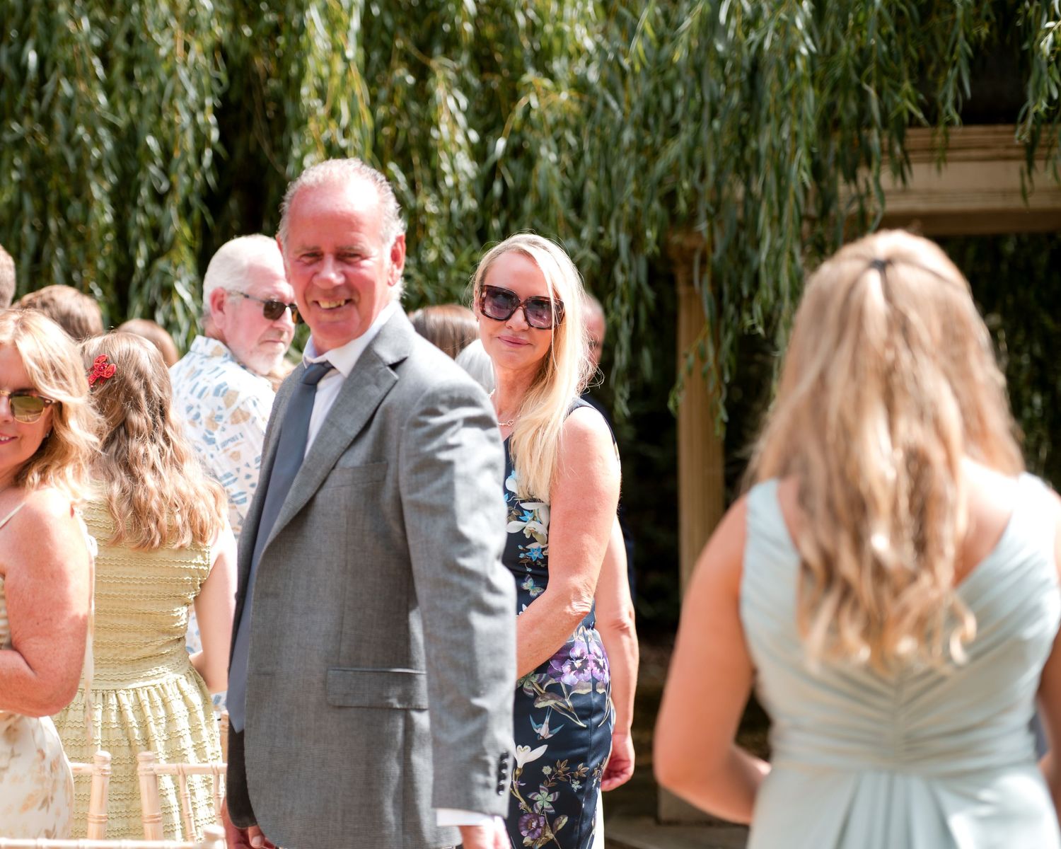 Man in gray suit smiling at outdoor wedding gathering with guests and willow trees in background.
