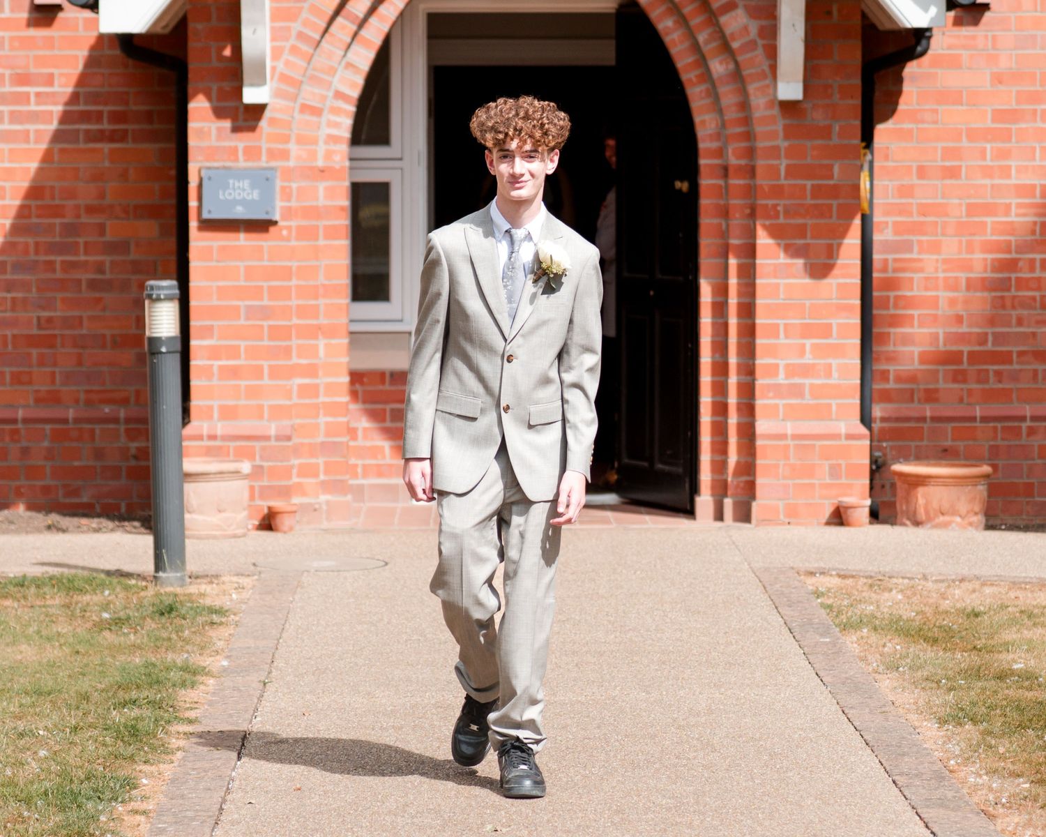 Young man in gray suit with boutonniere walking on pathway outside red brick church building.