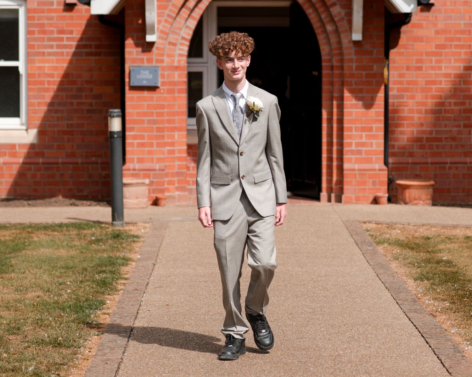 Young man in a light gray suit with boutonniere walking on a path outside a red brick building.