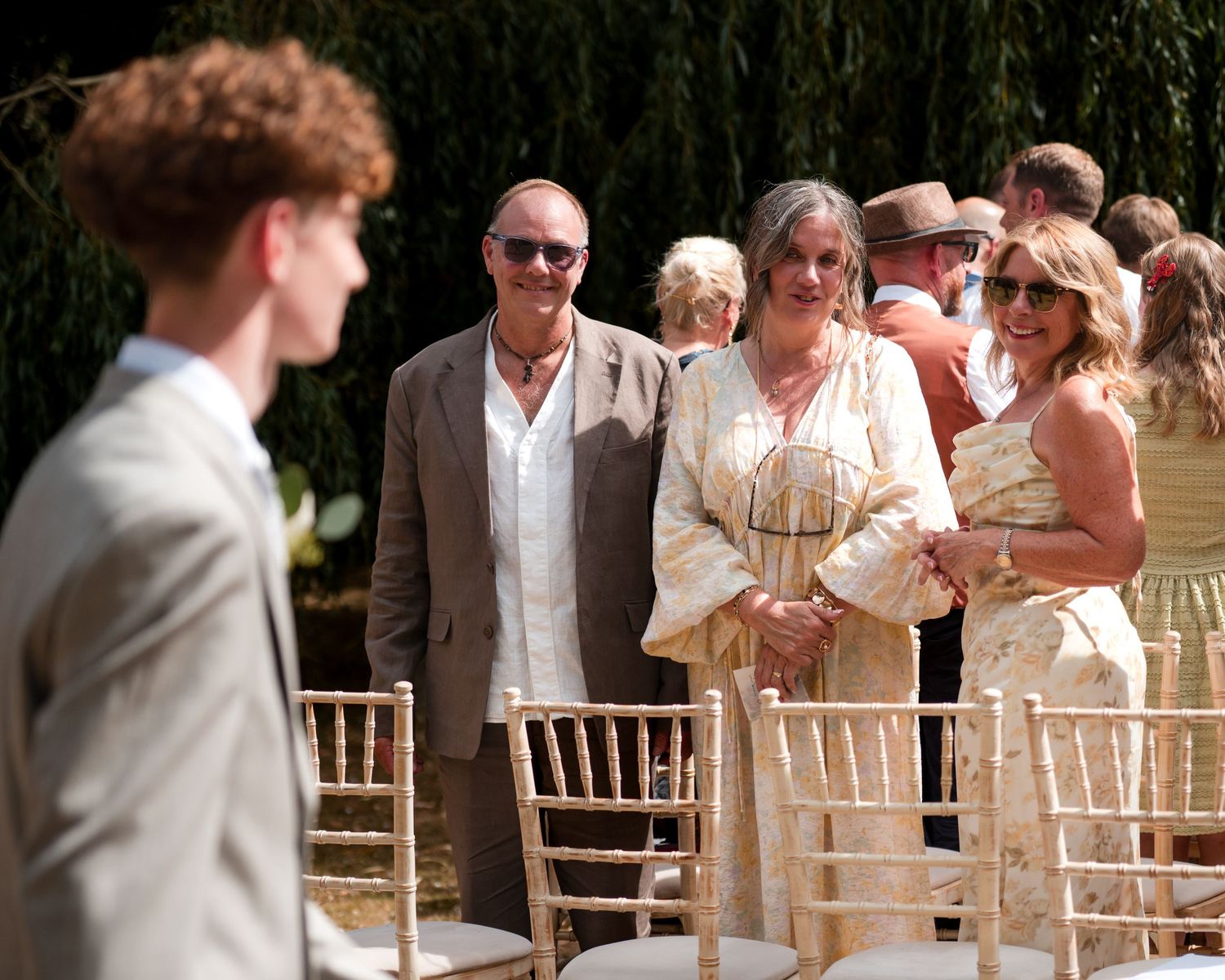 Guests smiling at an outdoor wedding ceremony seated on gold chiavari chairs in a sunny garden setting.