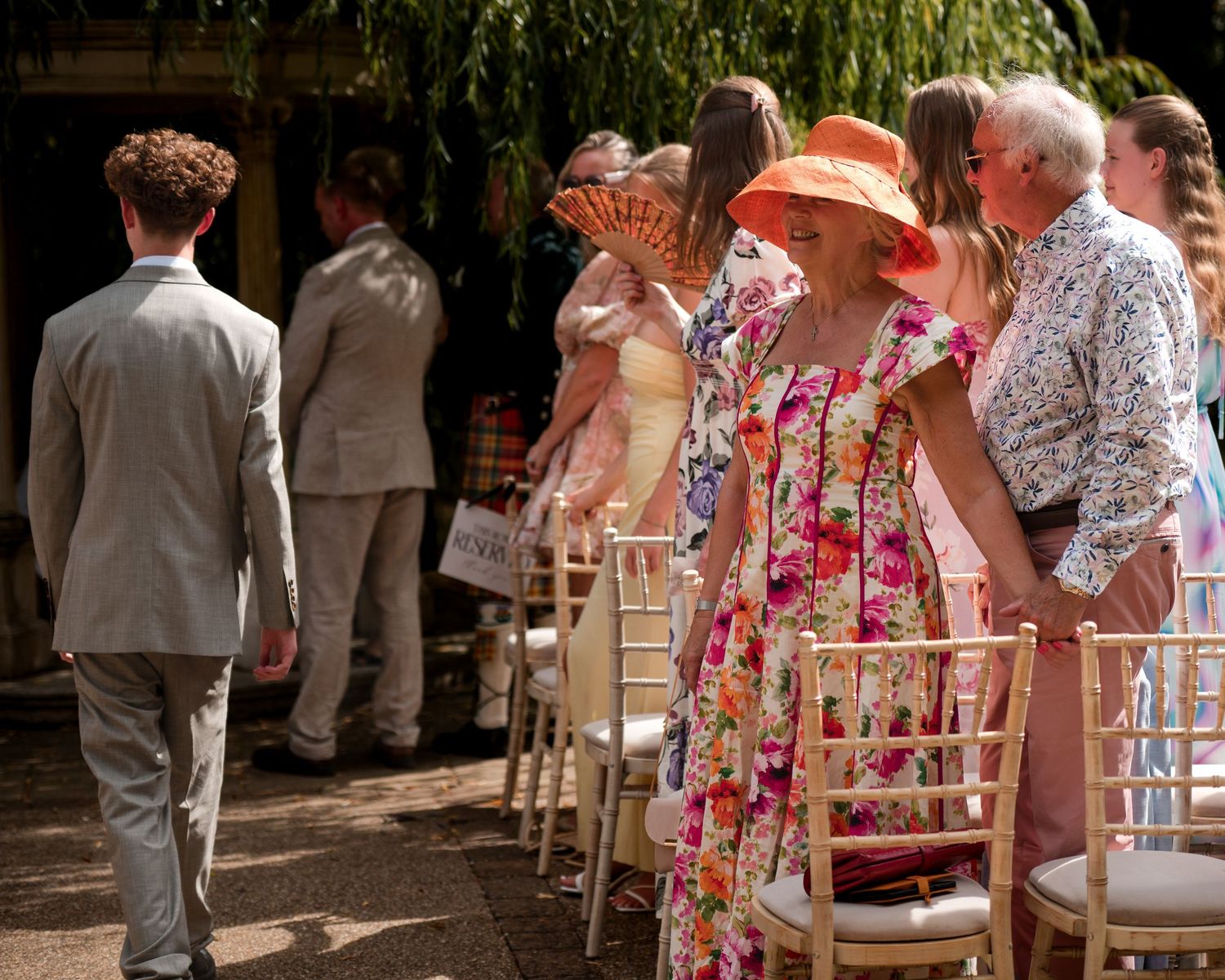 Elegantly dressed wedding guests in floral attire and orange hat stand beside chairs at an outdoor summer ceremony.