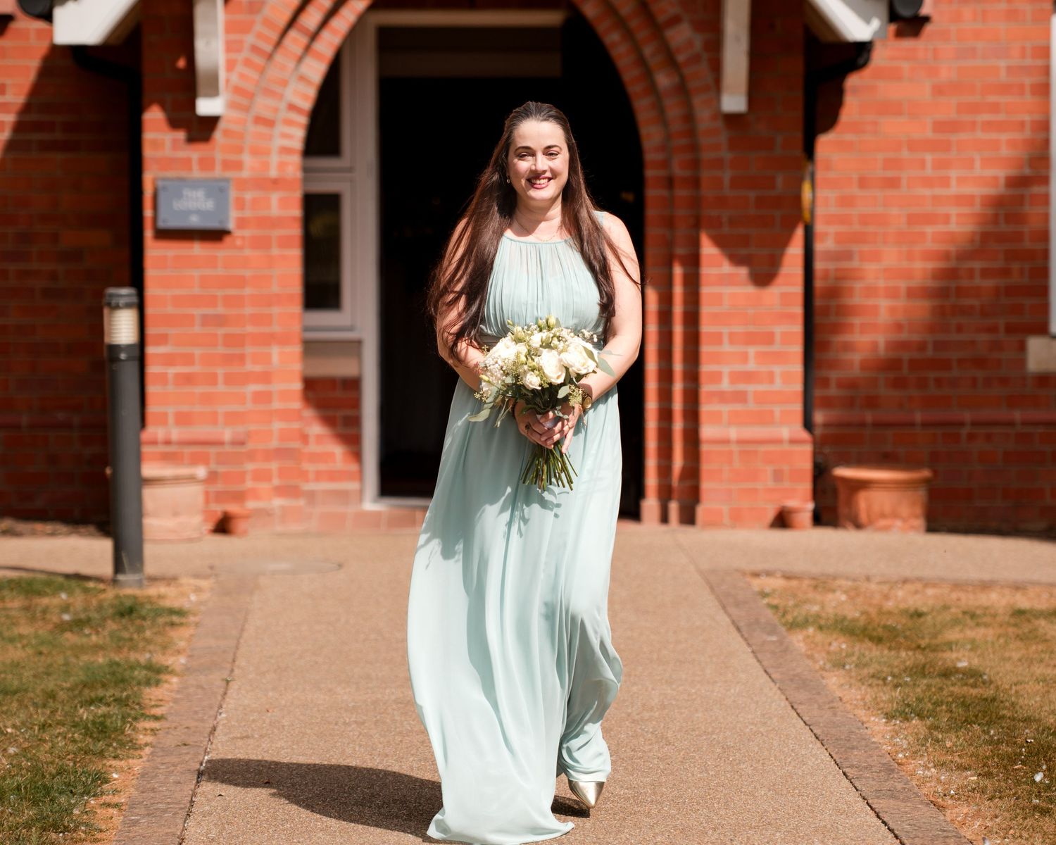 Bridesmaid in mint green gown holding bouquet walks toward camera outside brick church on sunny day.