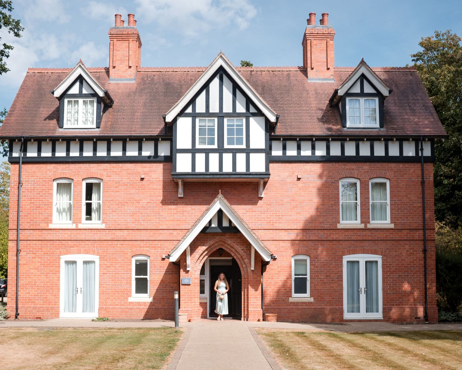 A grand red brick Tudor-style manor house with black and white timber framing and a person standing in the entrance doorway.