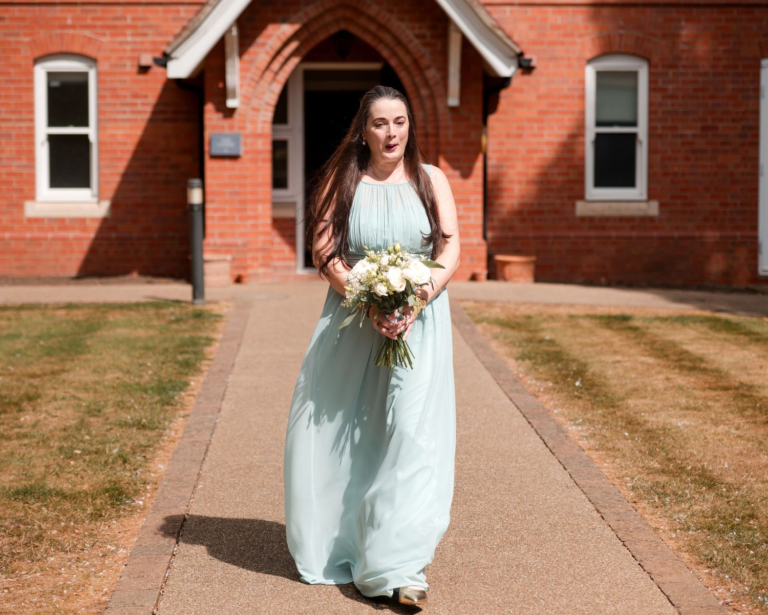 Bridesmaid in mint green gown holding white bouquet walks path outside red brick church on sunny day.