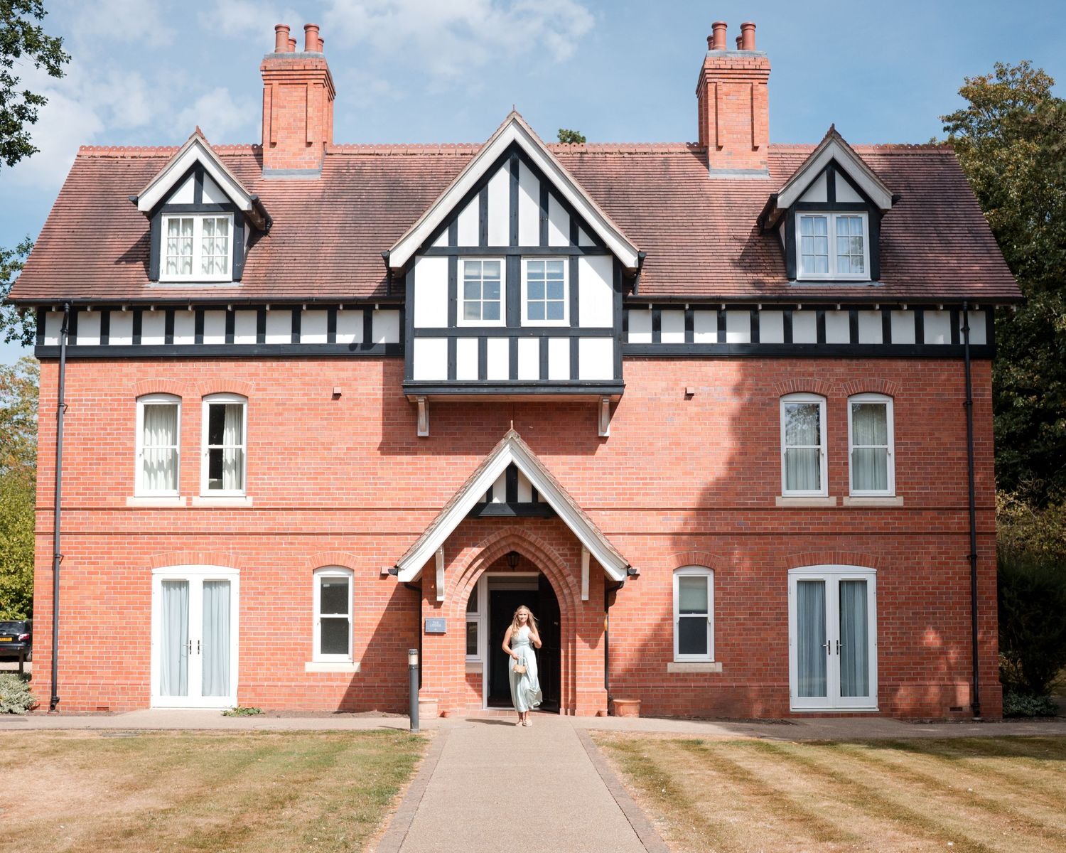 A grand red brick Tudor-style manor house with black and white timber framing and a person standing at the entrance.
