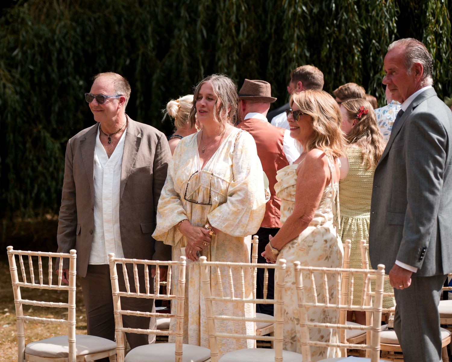 Guests in elegant attire stand beside chiavari chairs at an outdoor wedding ceremony surrounded by willow trees.