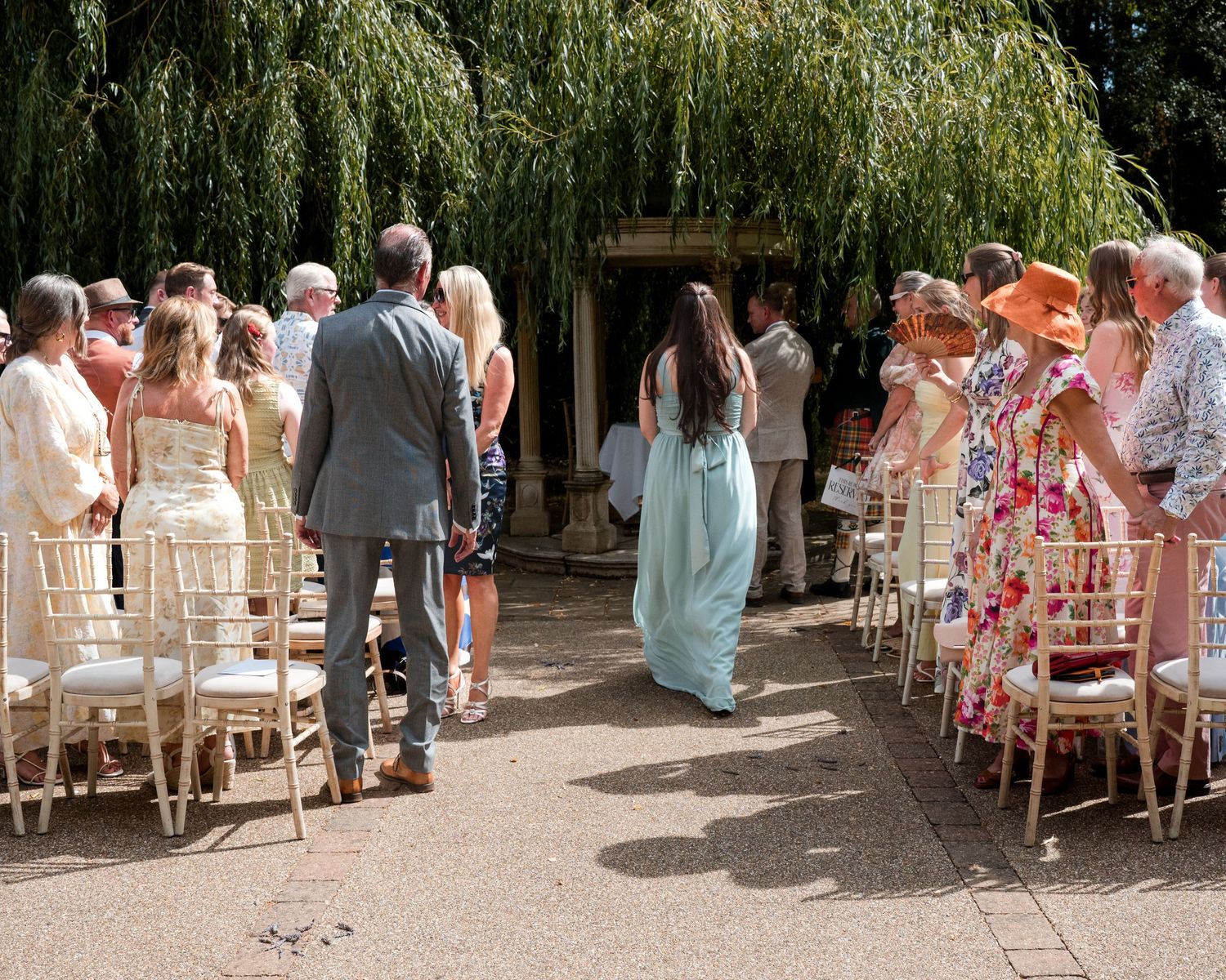 Outdoor wedding ceremony with guests seated on gold chairs along an aisle beneath a weeping willow tree.