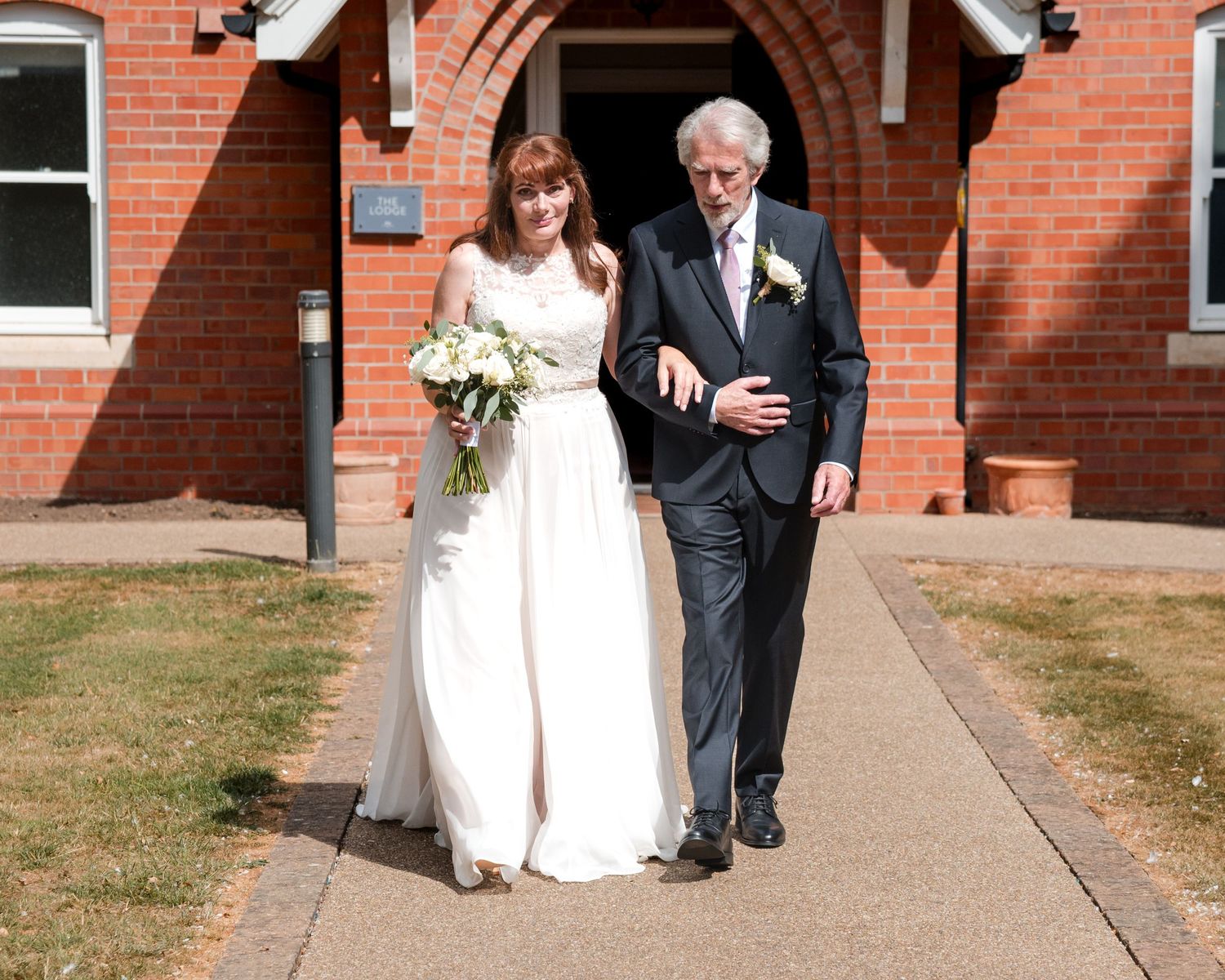 Bride in white gown walks with older gentleman in dark suit outside red brick church on sunny wedding day.