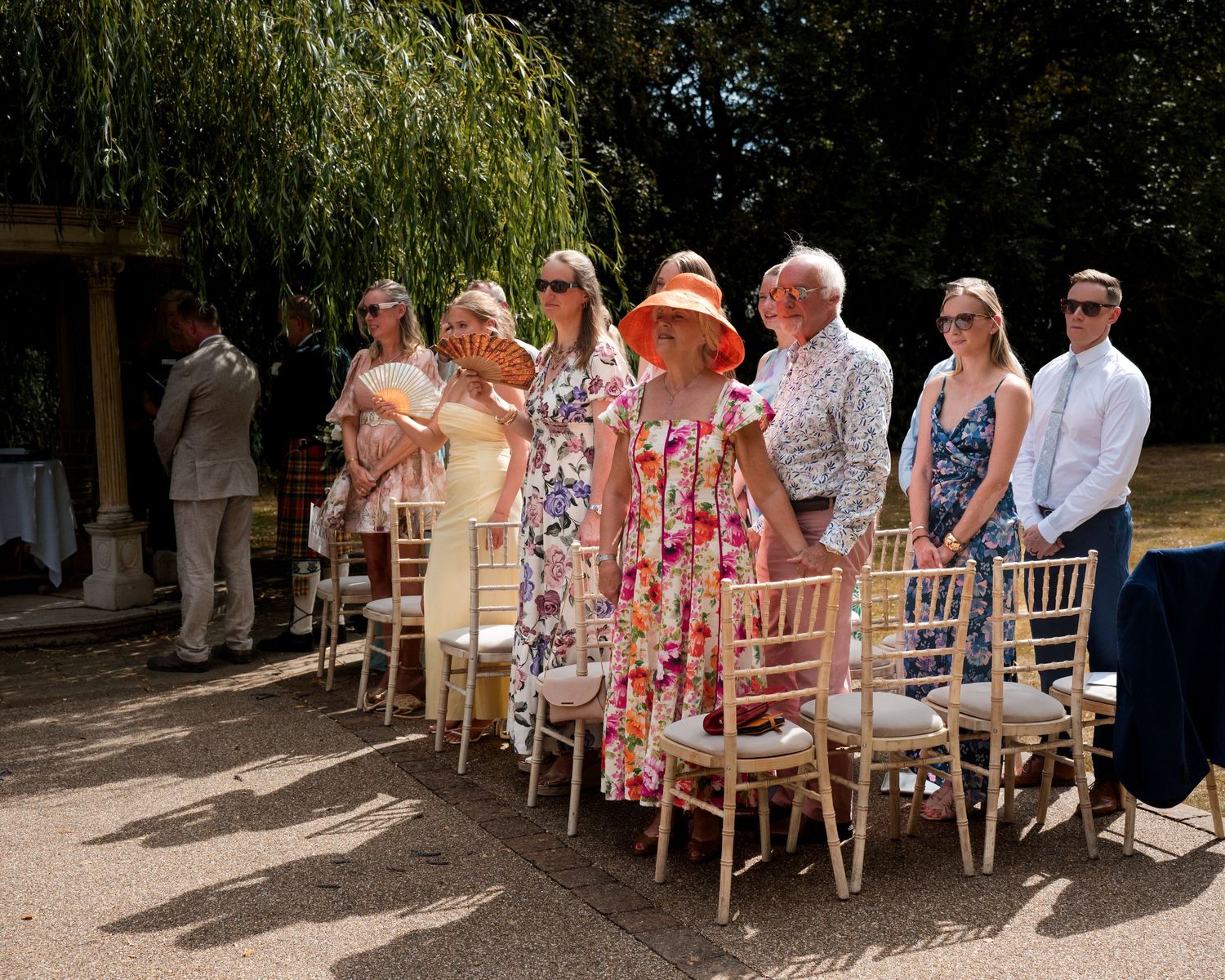 Guests in summer attire stand beside gold chiavari chairs at an outdoor garden wedding ceremony.