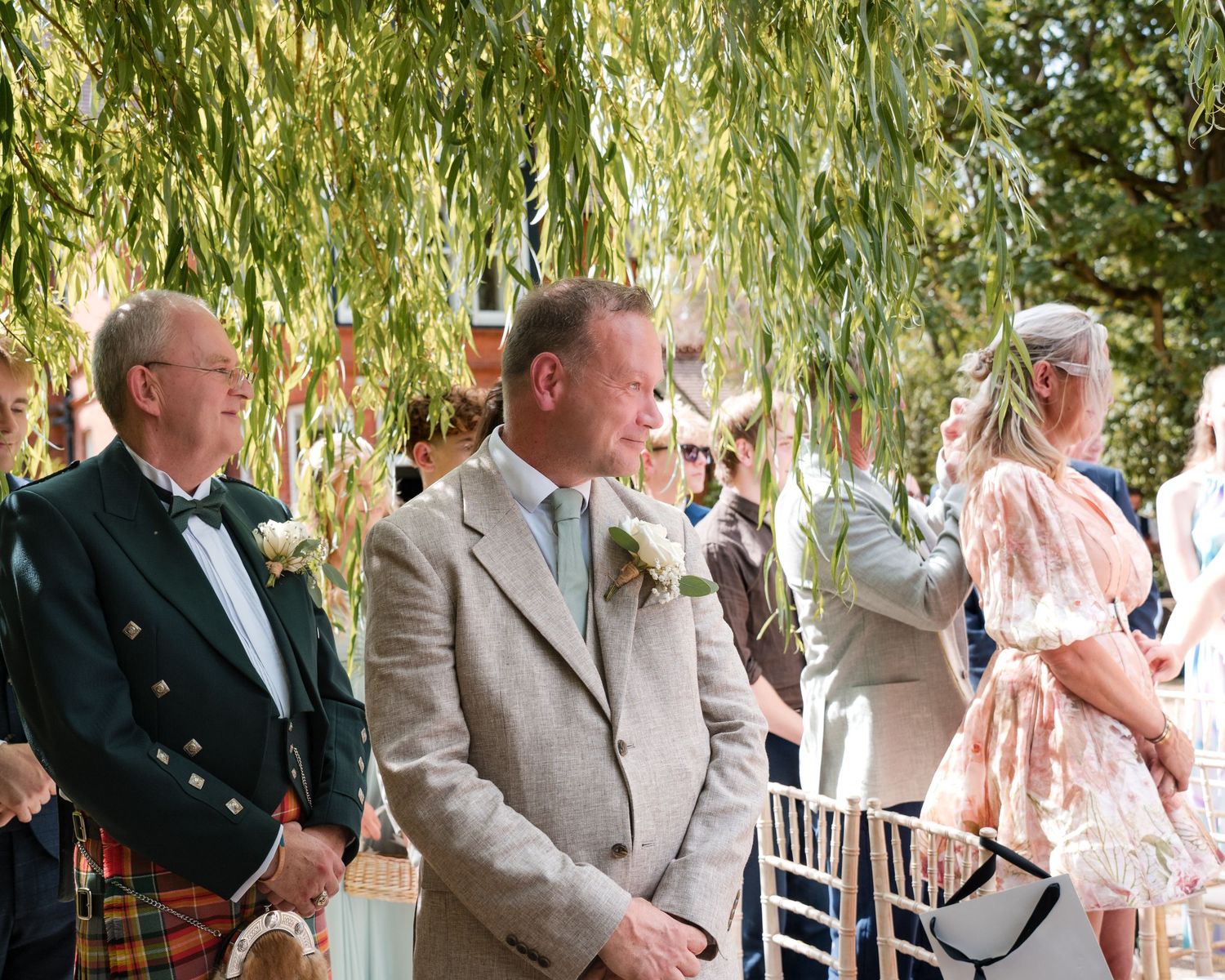 Two men in formal attire stand outdoors at a wedding ceremony beneath a weeping willow tree.