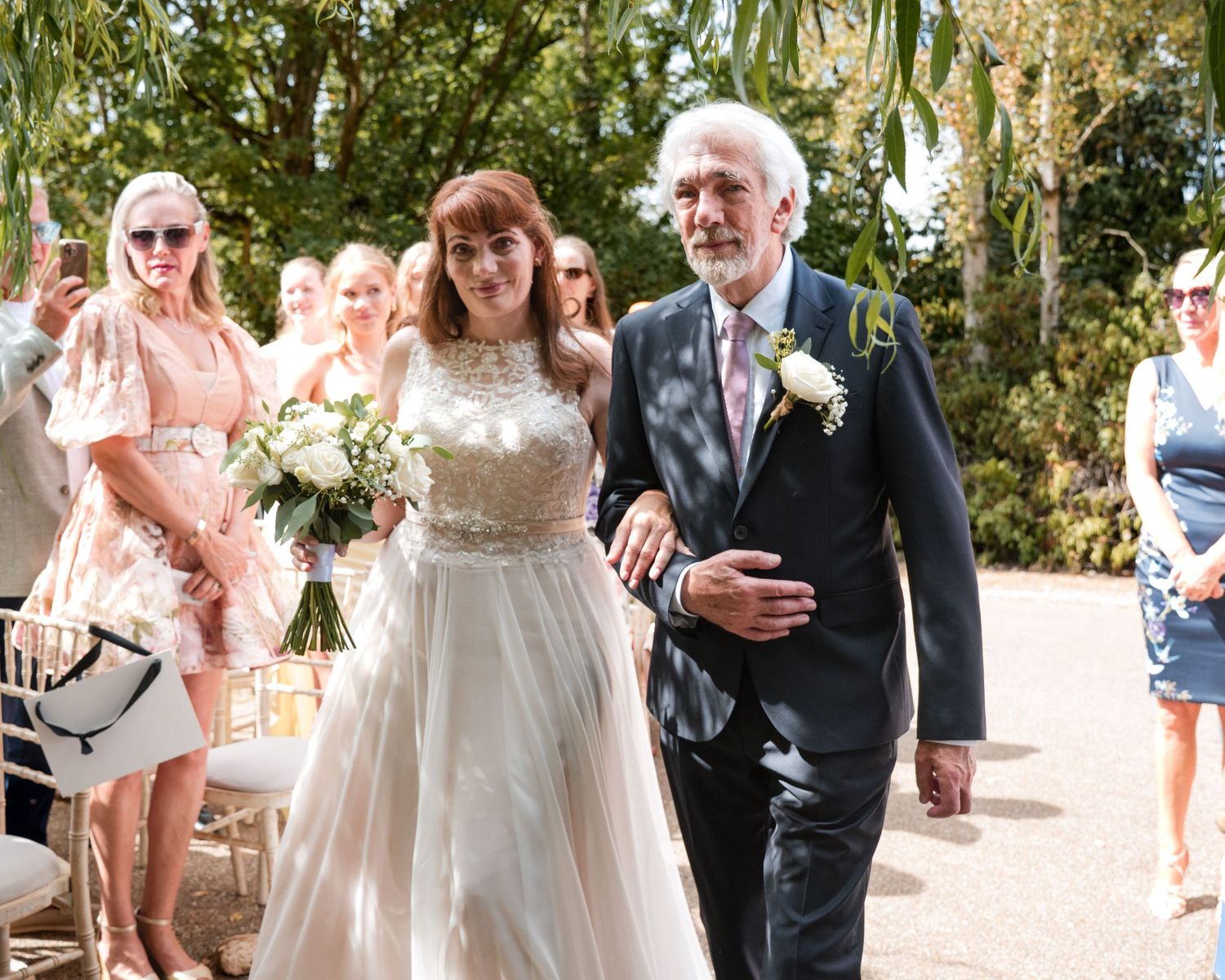 Bride in white gown walks down outdoor aisle with older gentleman, holding white bouquet, guests watching in sunny garden.