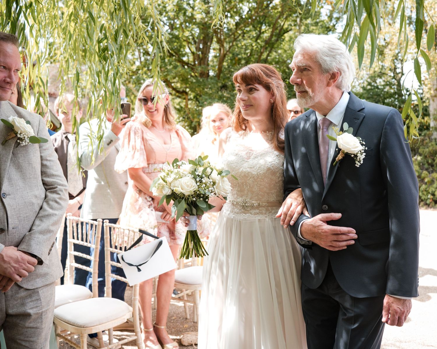Bride walks down the aisle with her father at an outdoor wedding ceremony under a willow tree.