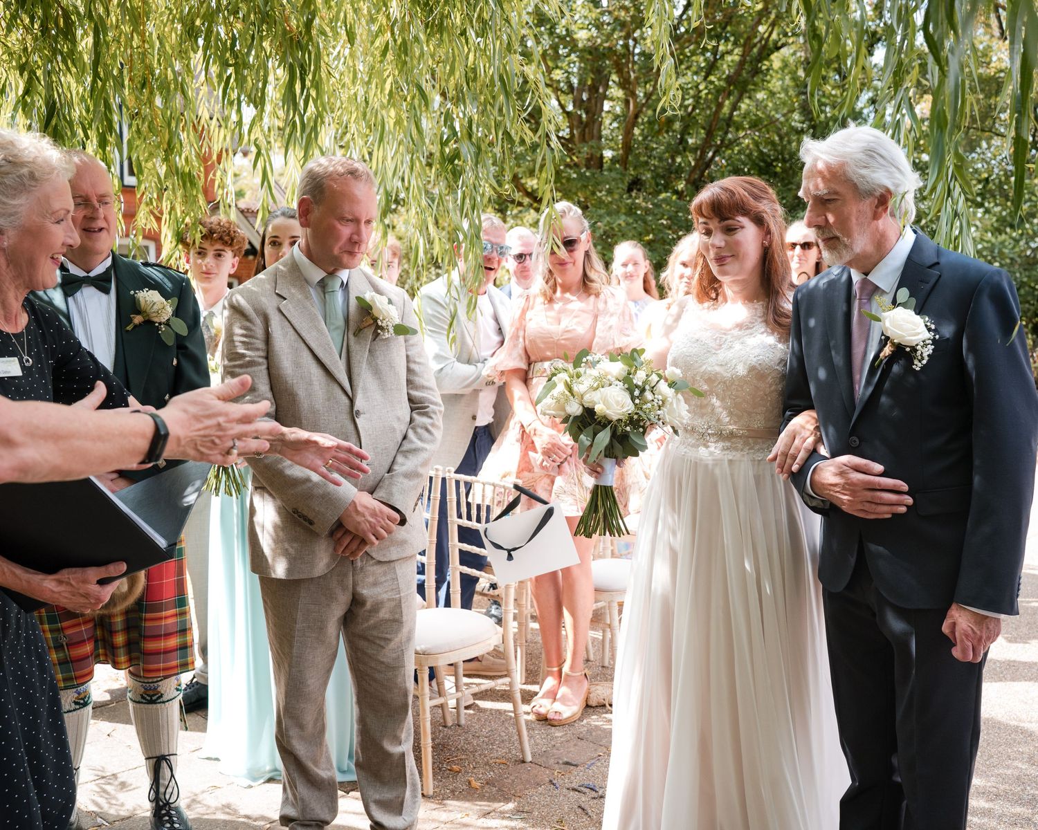 Bride walks down outdoor aisle with escort, groom waiting, guests seated among willow trees at sunny garden wedding.