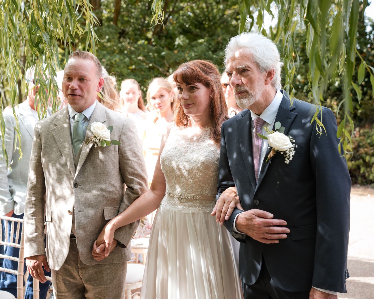 Bride in white gown walks down outdoor aisle with groom in tan suit and older man in navy blazer.