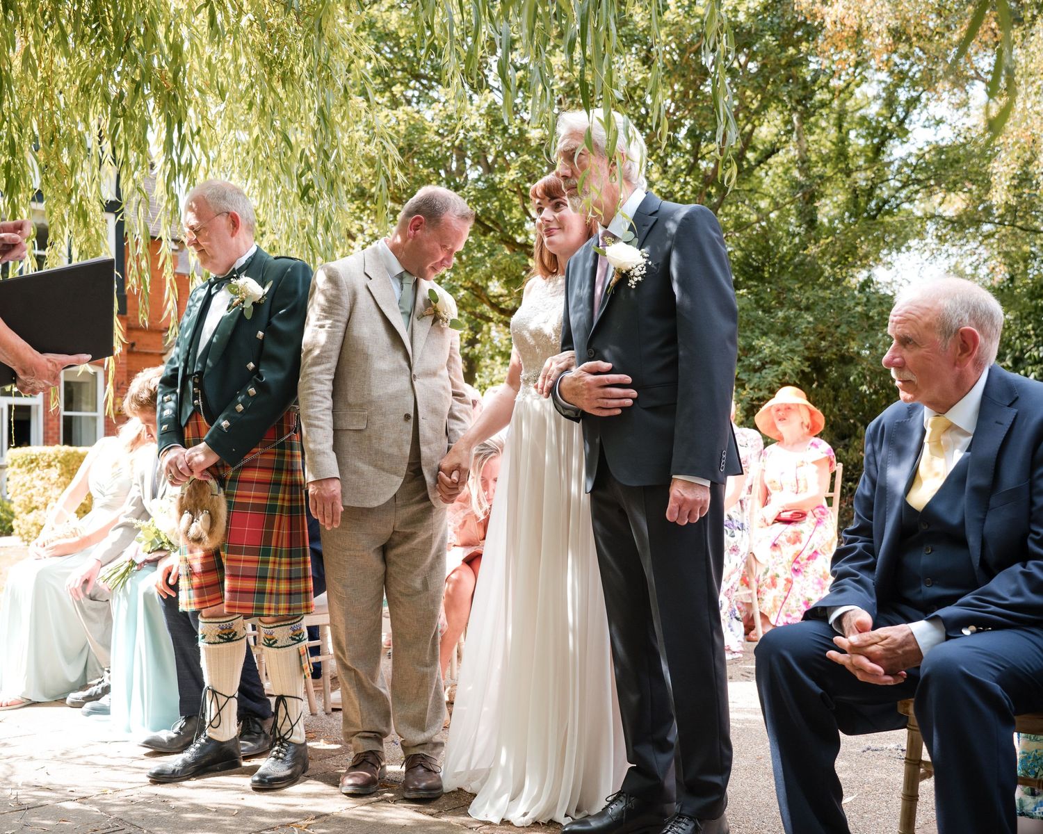 Outdoor wedding ceremony with bride and groom standing with guests, man in tartan kilt, under leafy trees.