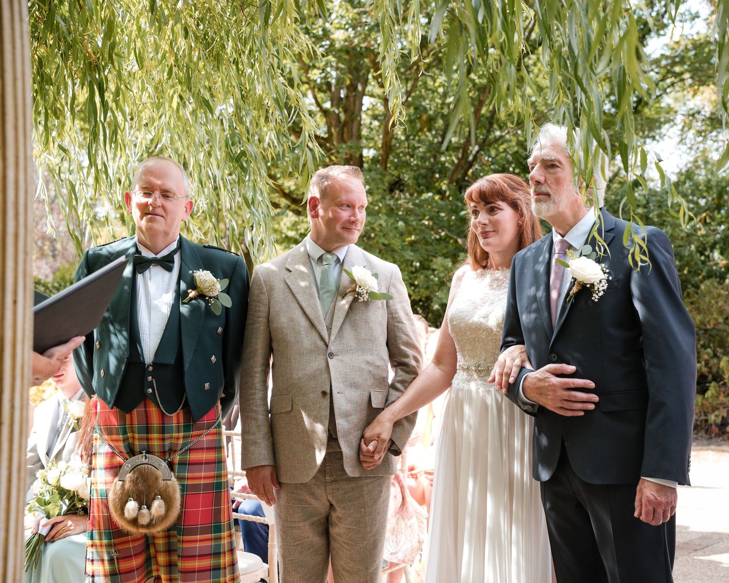 Four people pose outdoors at a wedding, including a man in a tartan kilt, a bride in white, and two suited gentlemen.
