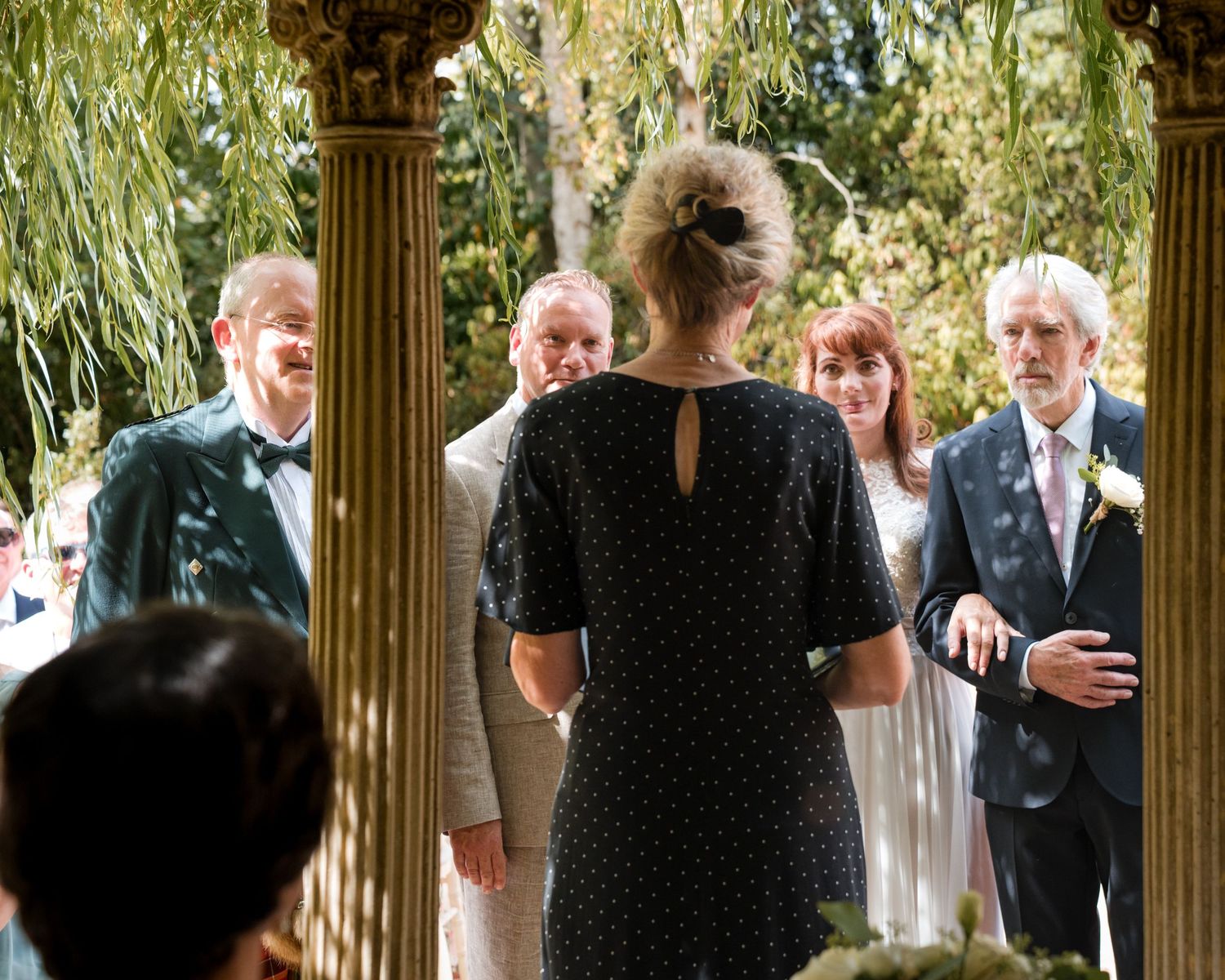 Outdoor wedding ceremony under classical columns with guests gathered around a woman in a black polka dot dress.