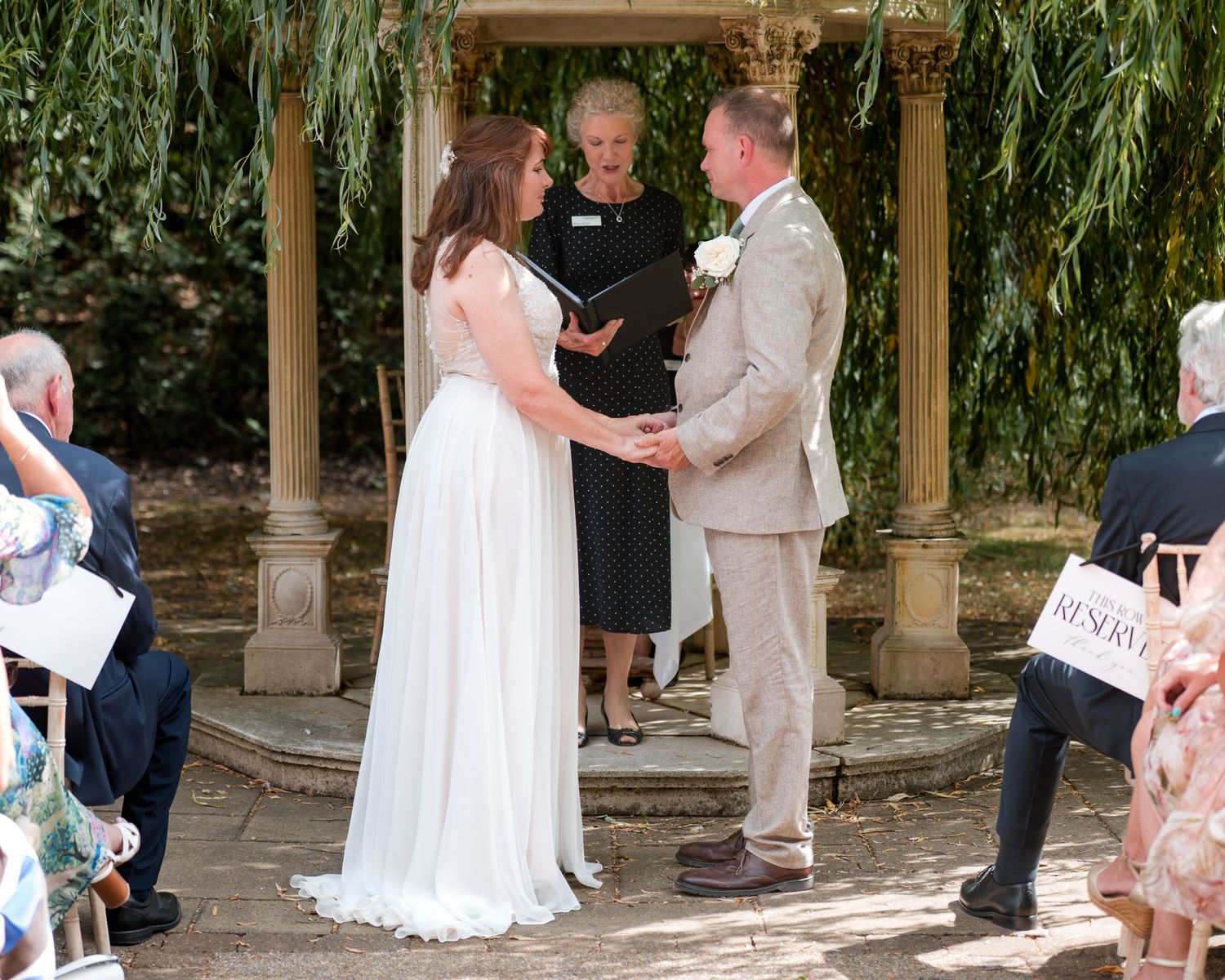 Couple exchanging vows at outdoor wedding ceremony under pergola with officiant, surrounded by lush greenery.