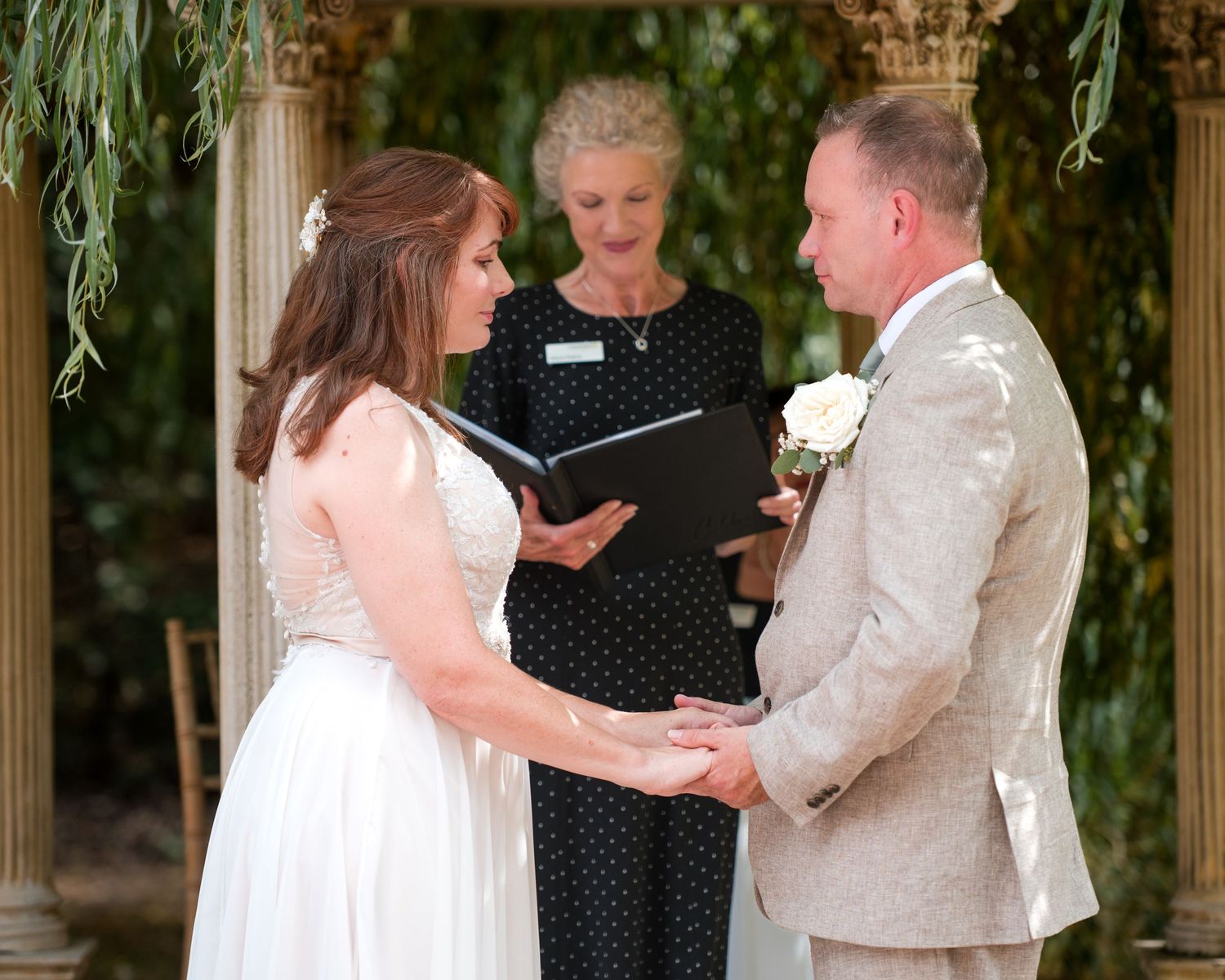 Bride and groom holding hands during outdoor wedding ceremony with officiant reading from book under floral arch.