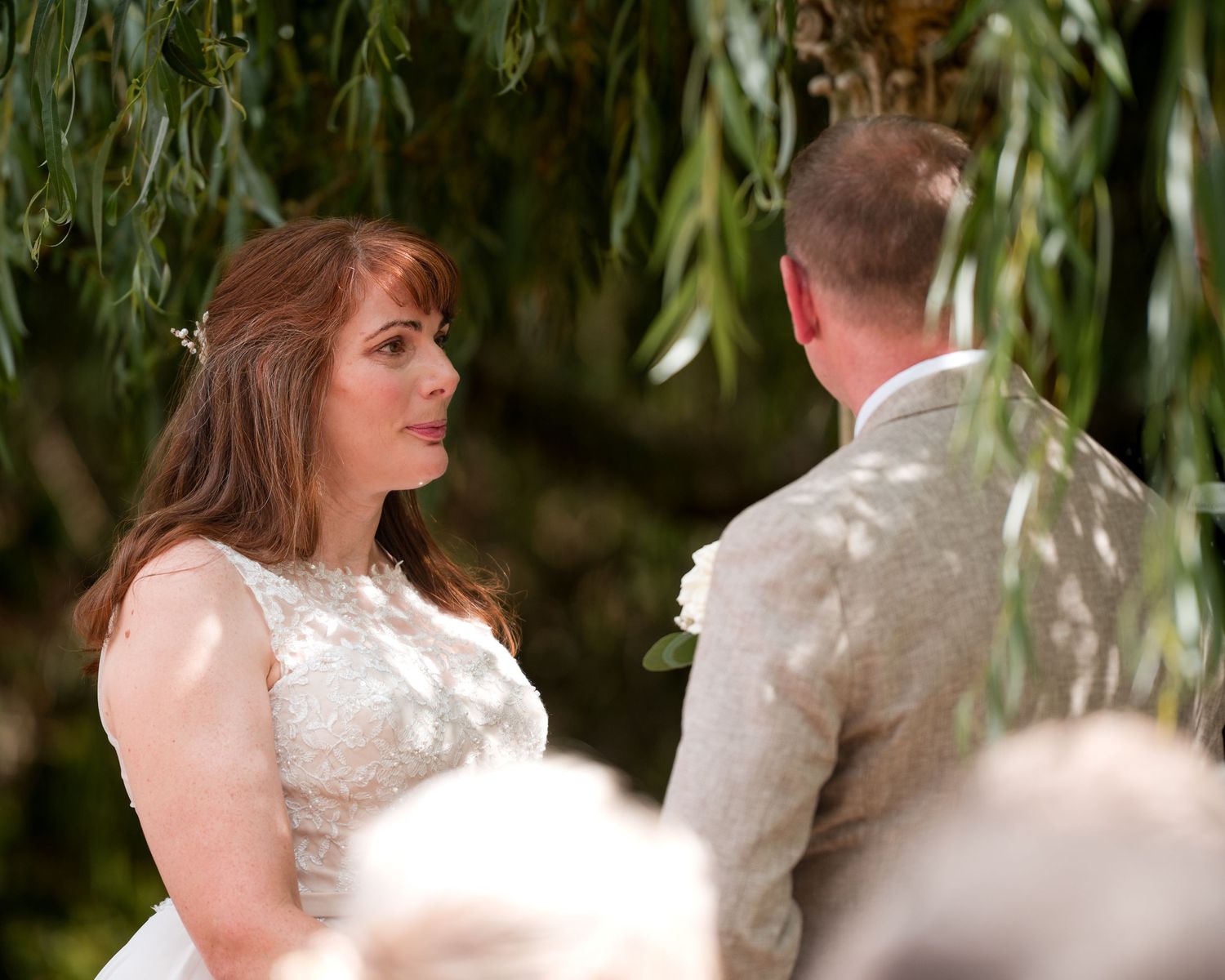 Bride in lace wedding dress exchanging vows with groom in tan suit outdoors under willow tree.