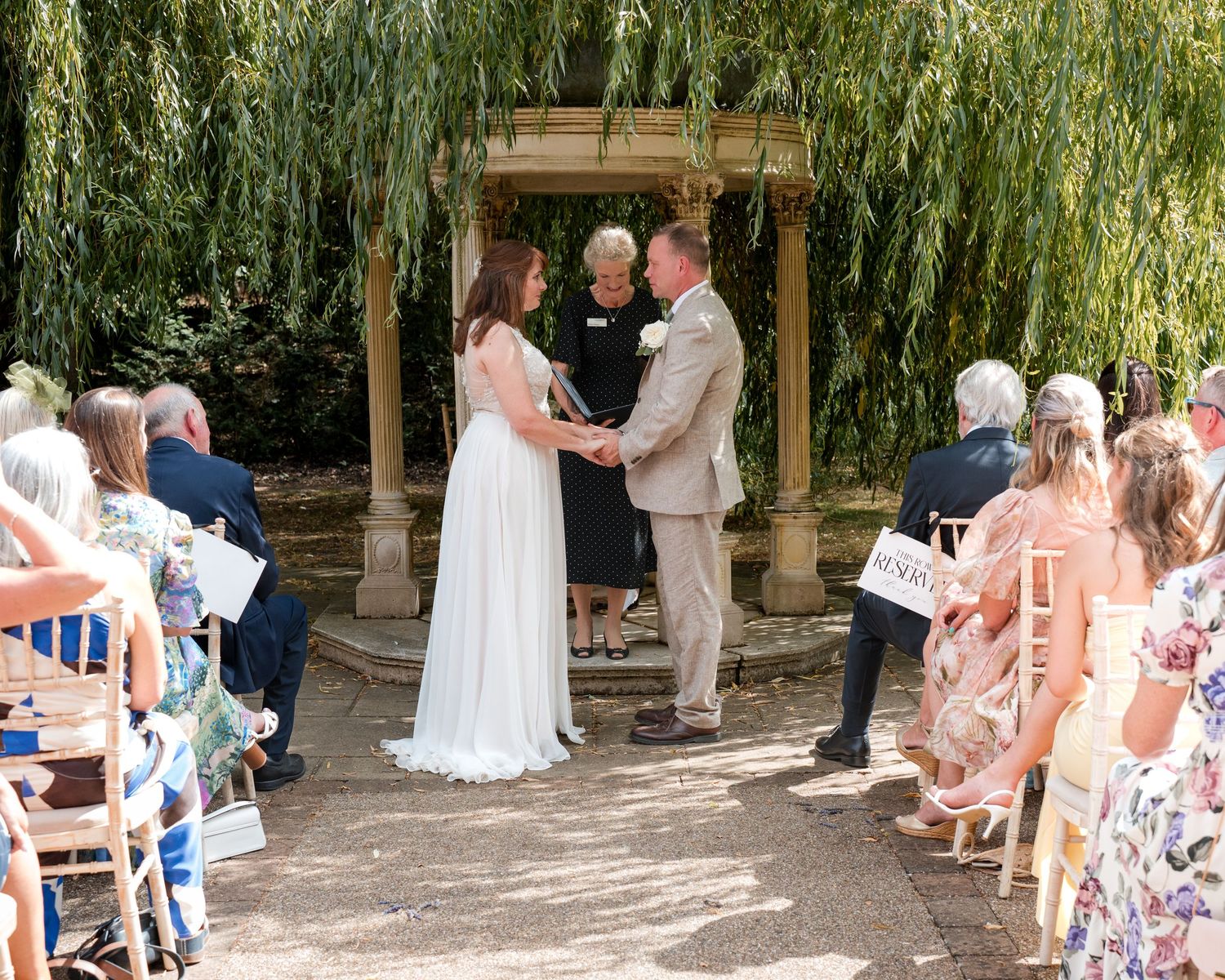 Outdoor wedding ceremony under a weeping willow tree, couple exchanging vows before officiant with seated guests.