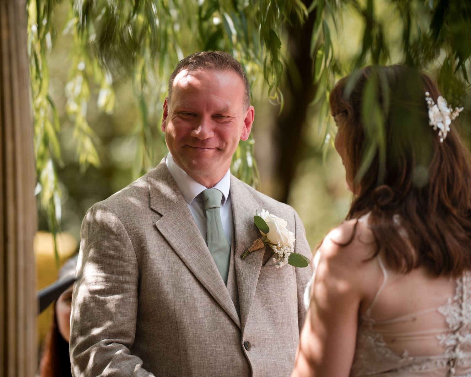 Groom smiling warmly at bride during outdoor wedding ceremony under willow tree, wearing gray suit with floral boutonniere.