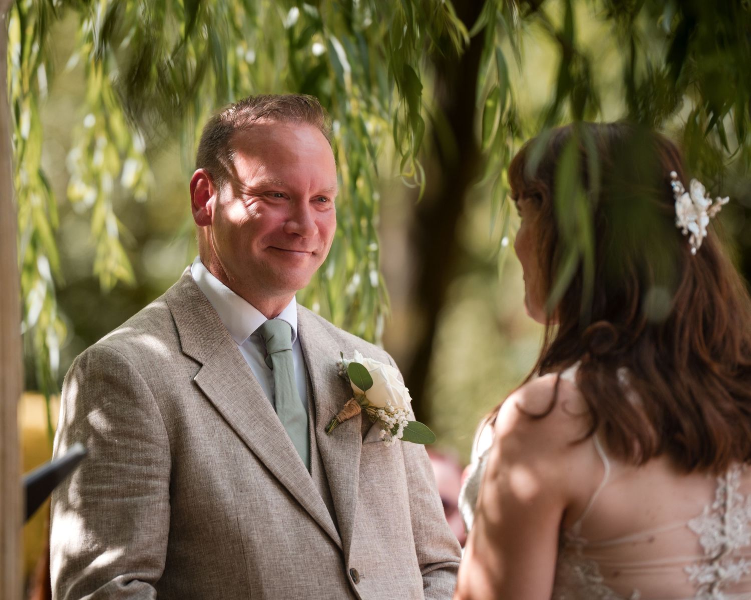 Groom in gray suit smiling at bride during outdoor wedding ceremony under weeping willow tree.