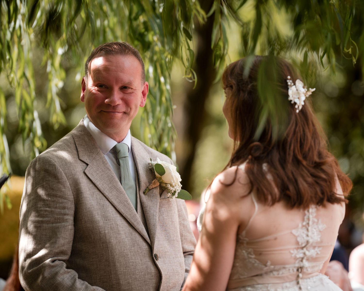 Groom in grey suit smiling at bride during outdoor wedding ceremony under weeping willow tree.