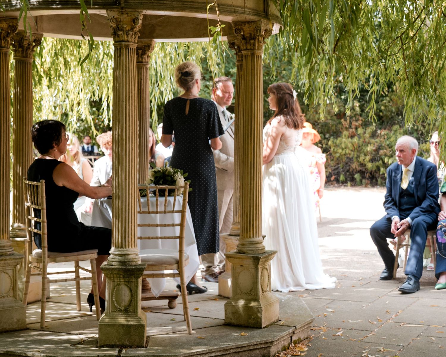 Outdoor wedding ceremony under a stone gazebo surrounded by weeping willow trees with guests seated nearby.