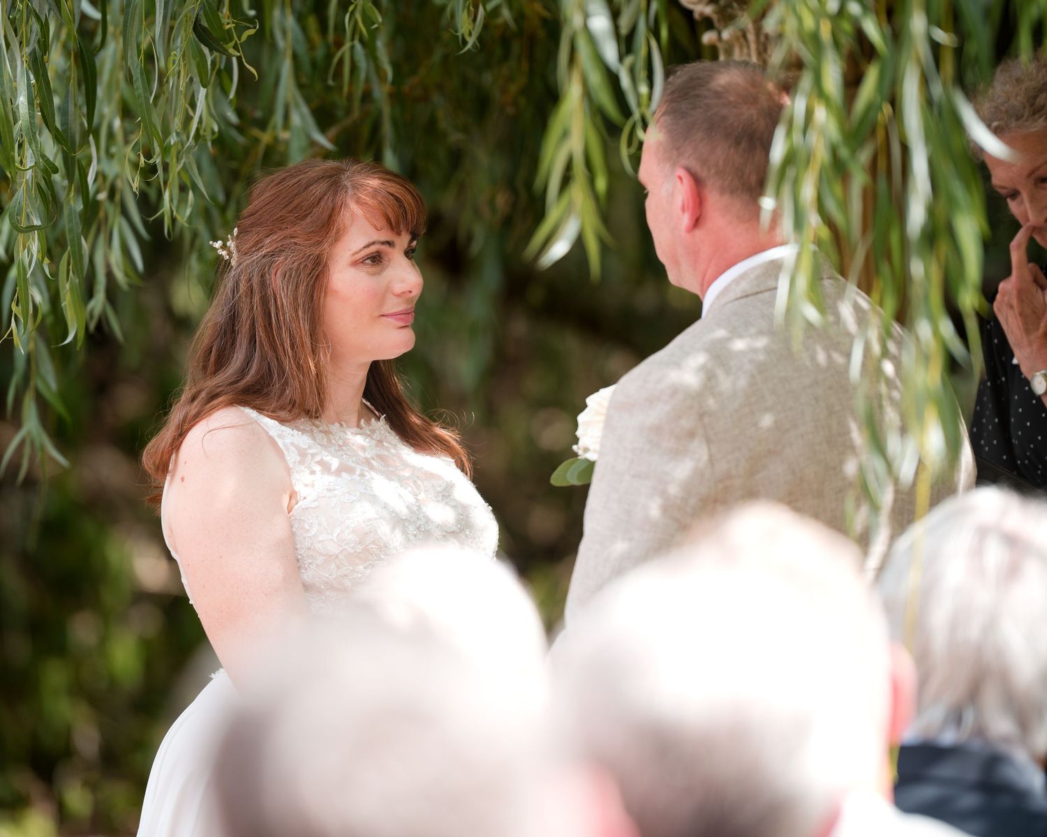 Bride in lace wedding dress exchanges vows with groom outdoors under weeping willow tree.