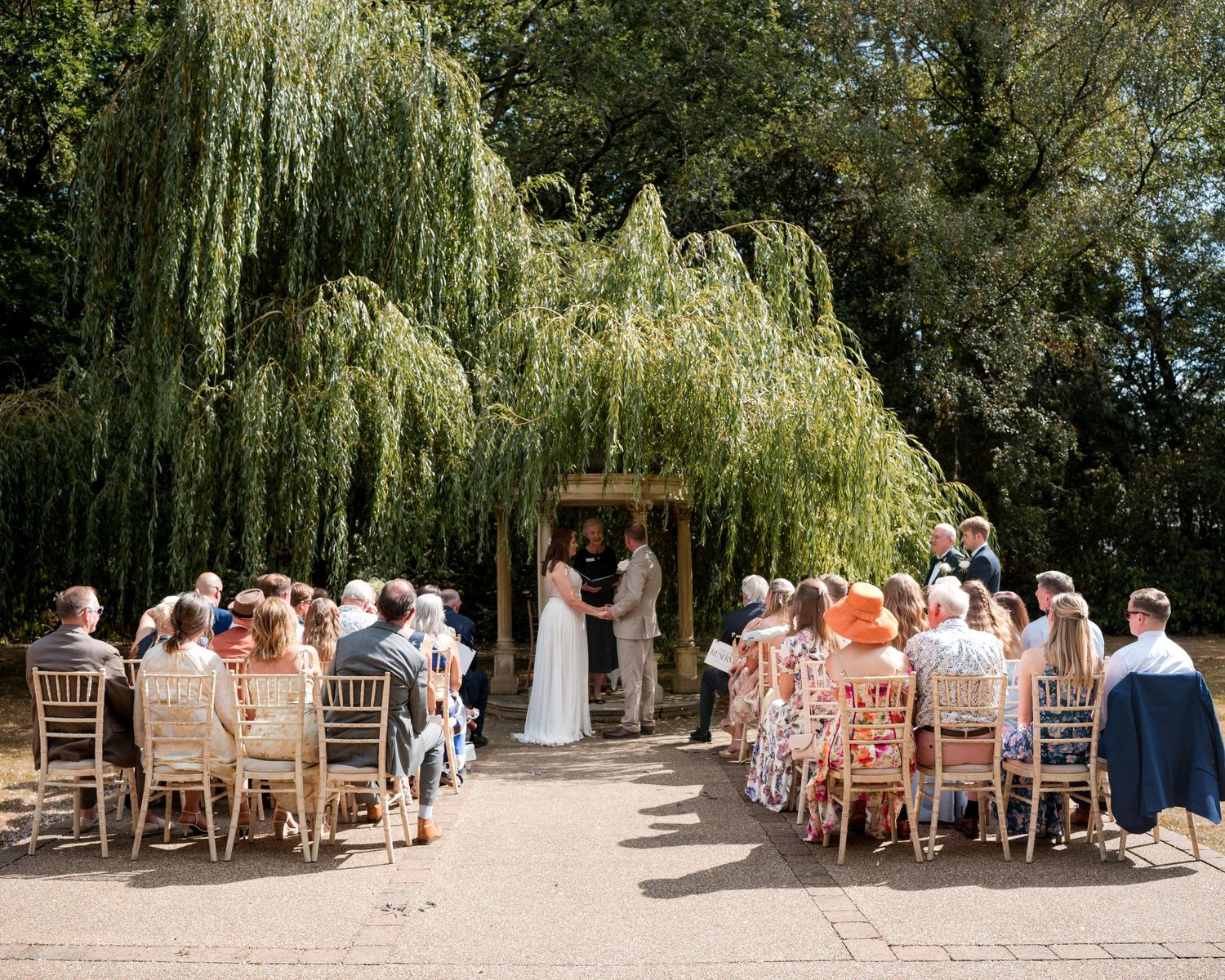 Outdoor wedding ceremony under a weeping willow tree with guests seated on wooden chairs in sunny garden.