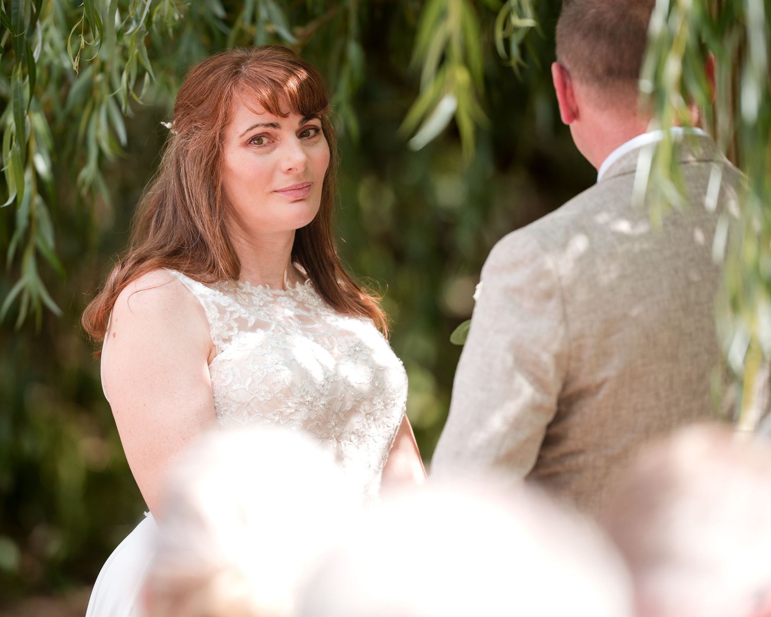 Bride in white lace gown gazes at groom during outdoor wedding ceremony under weeping willow tree.