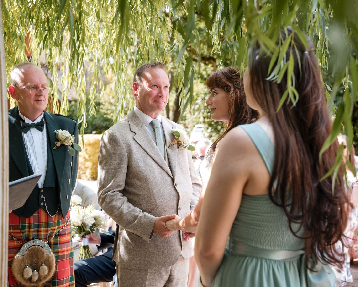 Groom in beige suit exchanges vows with bride in mint dress, officiant in Scottish kilt stands beside them outdoors.