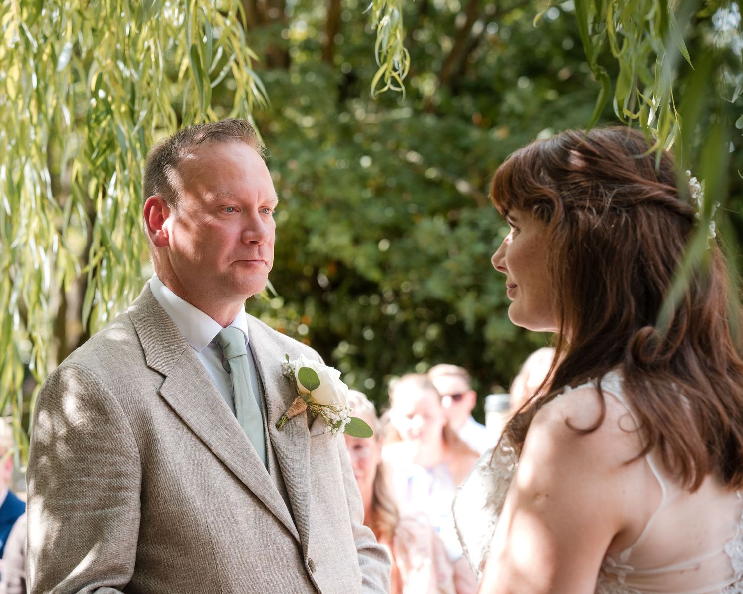 Couple exchanging vows at outdoor wedding ceremony under weeping willow tree, groom in gray suit with boutonniere.