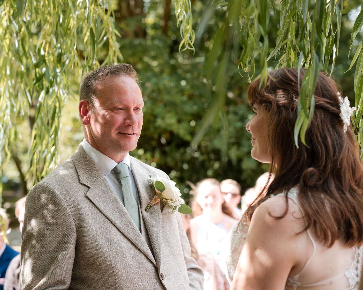 Couple exchanging vows at an outdoor wedding ceremony under weeping willow trees, groom in gray suit.