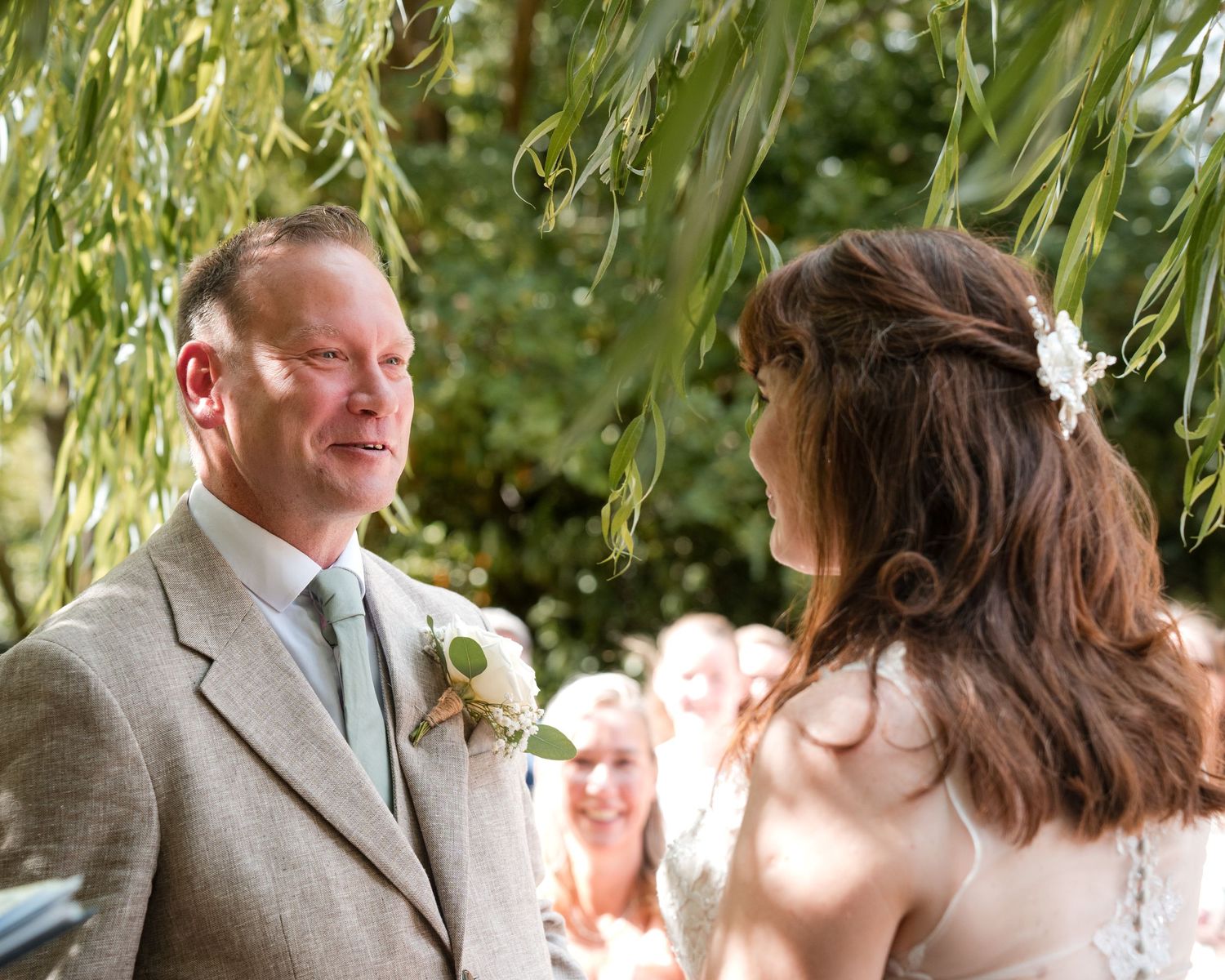 Bride and groom exchange vows outdoors under willow tree at elegant garden wedding ceremony.