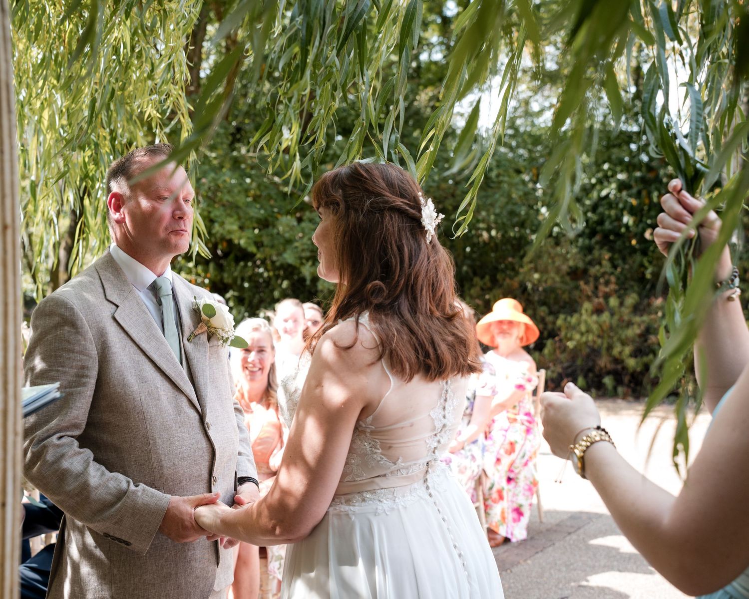 Bride and groom hold hands during outdoor wedding ceremony under weeping willow tree with guests in background.