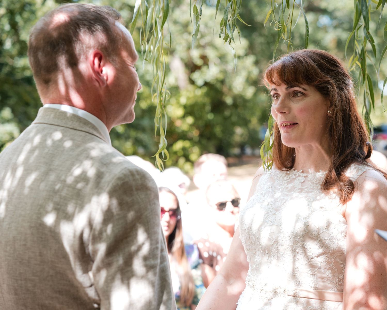 Bride and groom exchange vows outdoors under willow tree at sunny garden wedding ceremony.