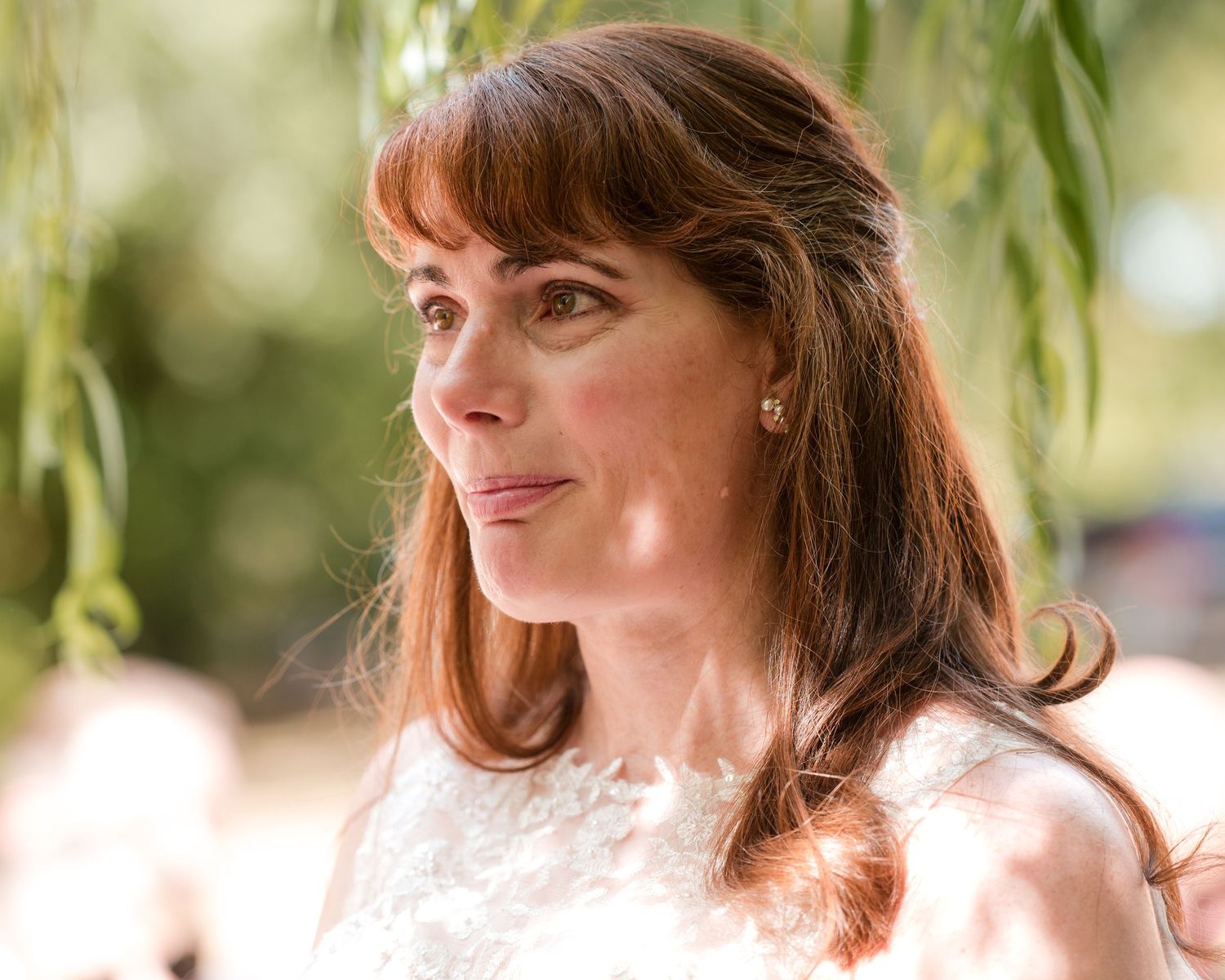 A woman with auburn hair and bangs wearing a white dress, gazing upward outdoors near a willow tree.