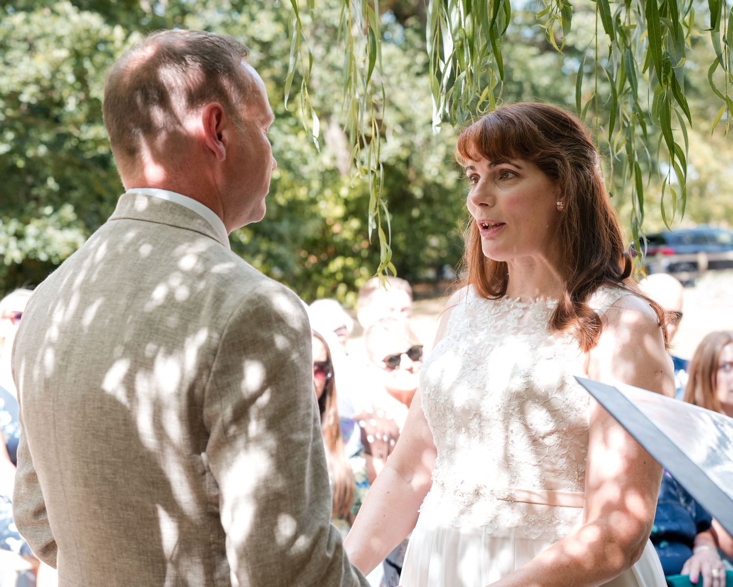 Bride and groom exchange vows outdoors under willow tree at sunny wedding ceremony with guests seated behind.