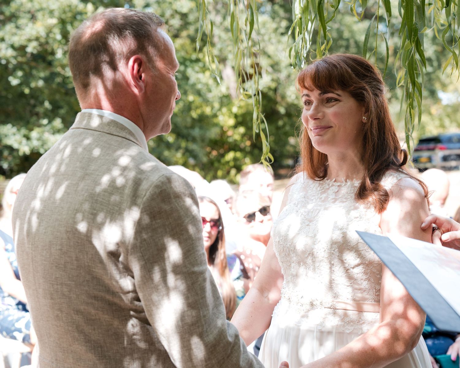 Bride and groom exchange vows outdoors under willow tree at sunny garden wedding ceremony.