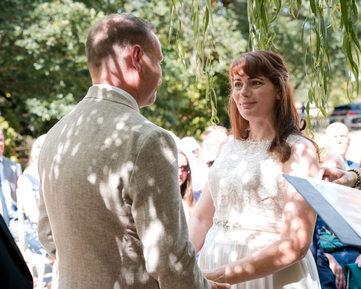 Bride and groom exchanging vows at outdoor wedding ceremony beneath sunlit willow tree.