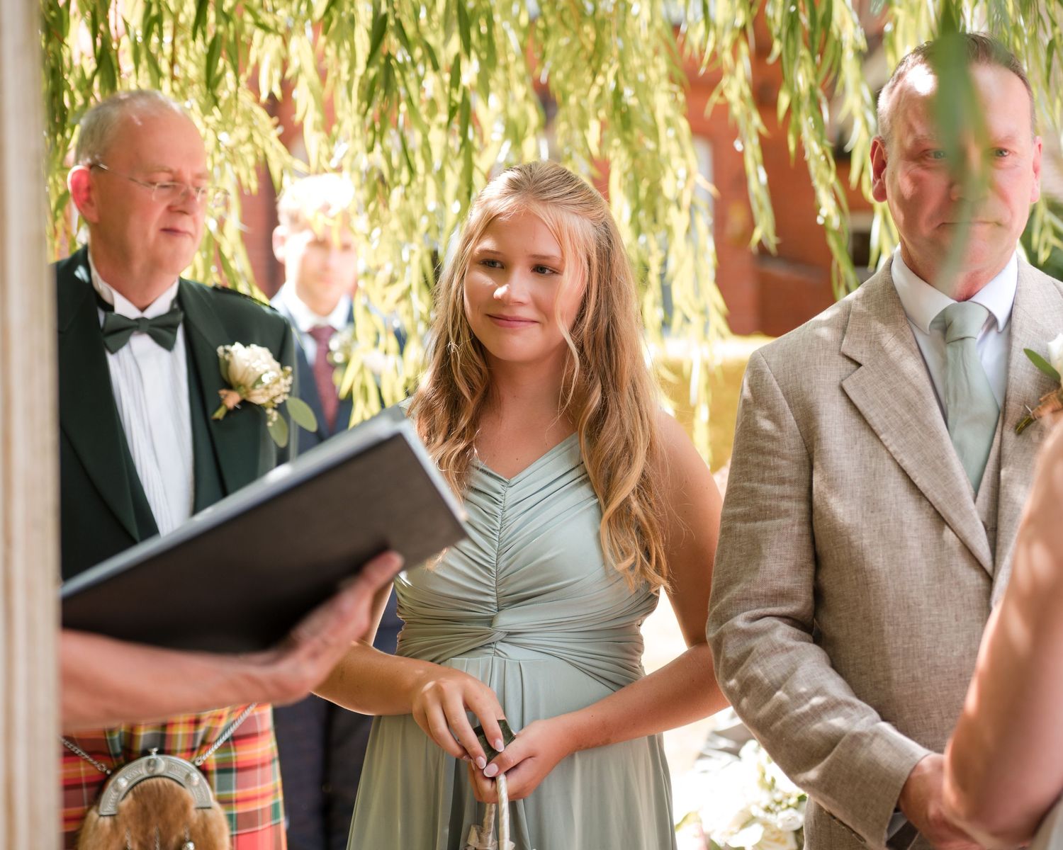 Smiling bridesmaid in sage green dress stands beside groom during outdoor wedding ceremony.