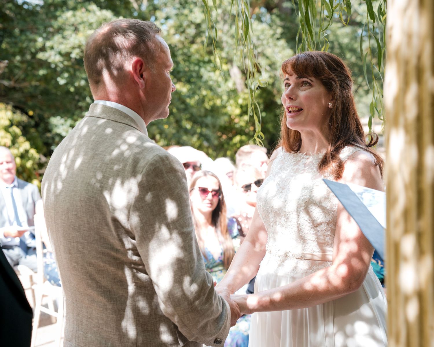 Couple exchanging vows at an outdoor wedding ceremony, bride in white dress, groom in grey suit, lush greenery background.