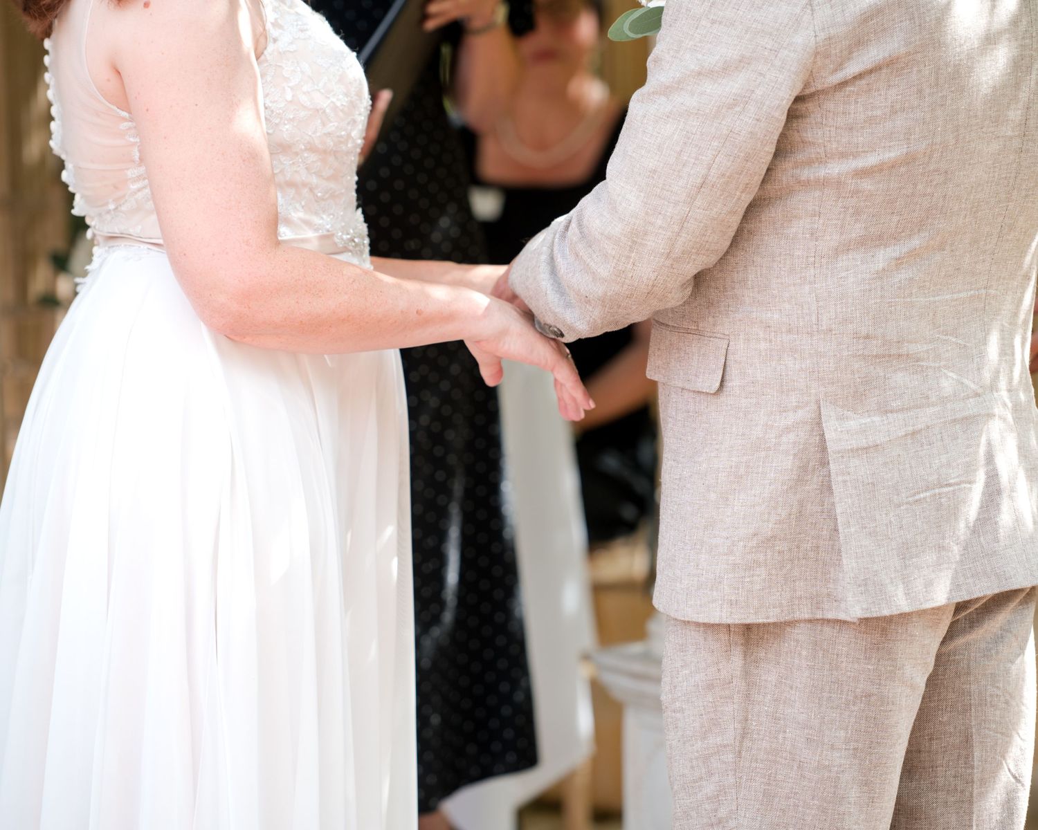 Bride and groom holding hands during wedding ceremony, bride in white gown, groom in beige suit.