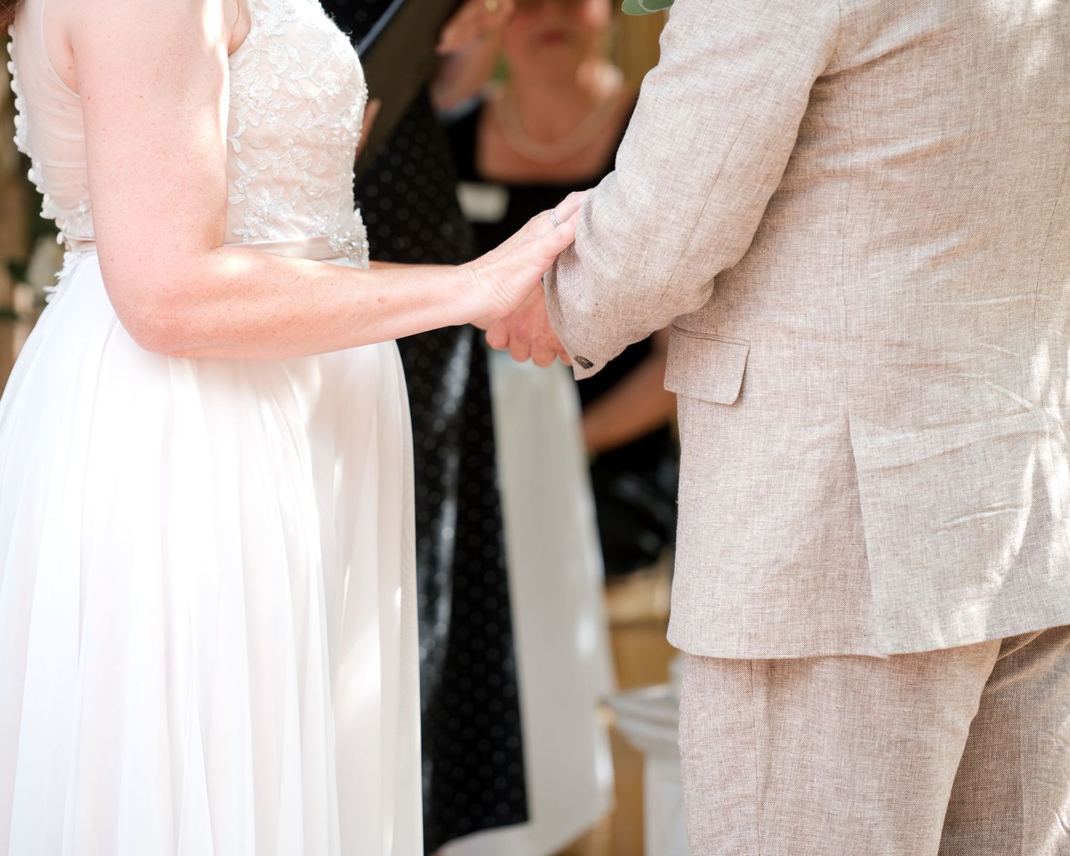 Bride and groom holding hands during outdoor wedding ceremony, bride in white gown, groom in beige suit.