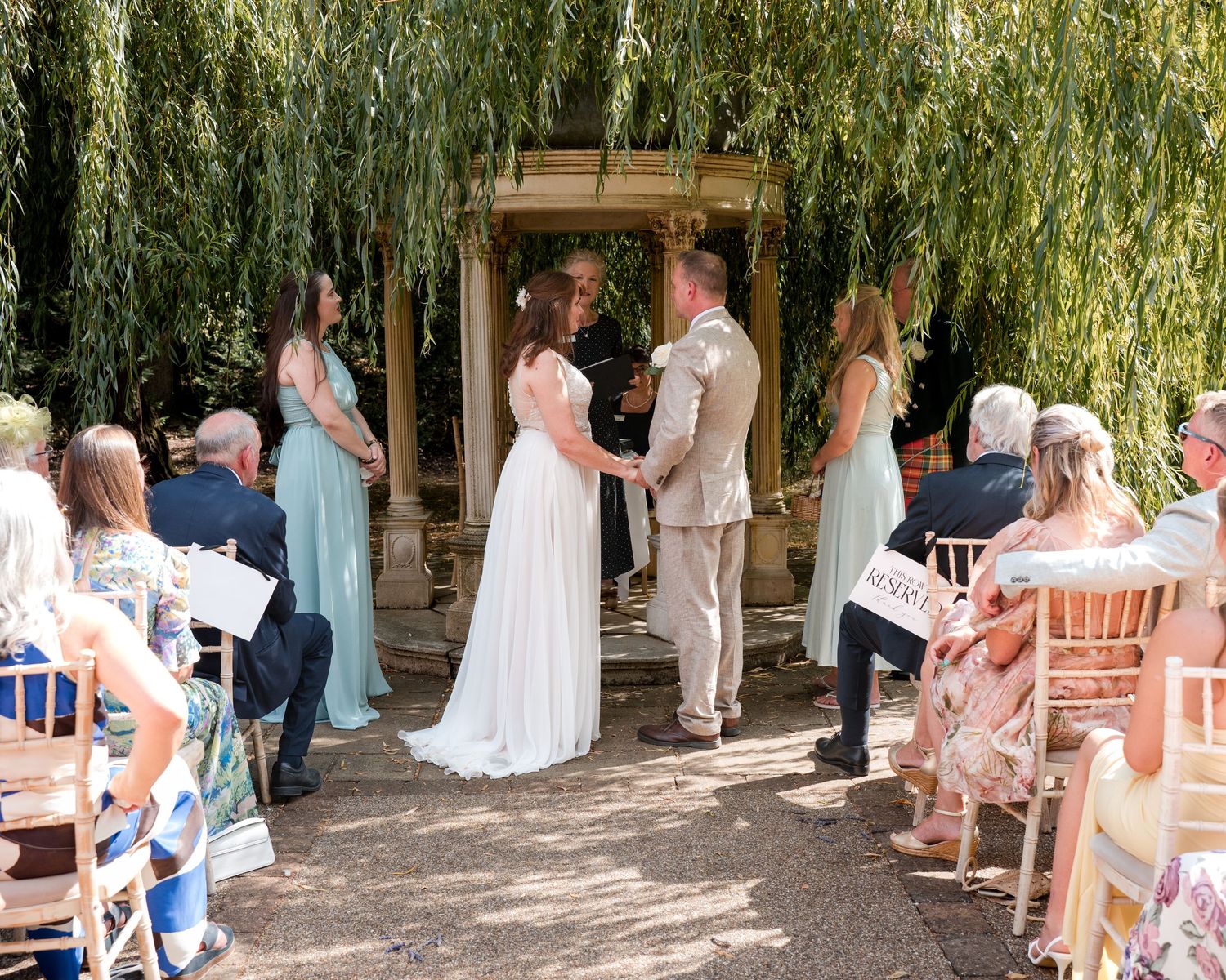 Outdoor wedding ceremony under a weeping willow tree with bride and groom exchanging vows before seated guests.