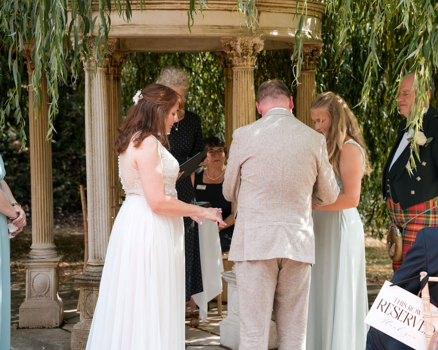 Bride and groom exchange vows at outdoor wedding ceremony under elegant pavilion with bridesmaid and officiant present.