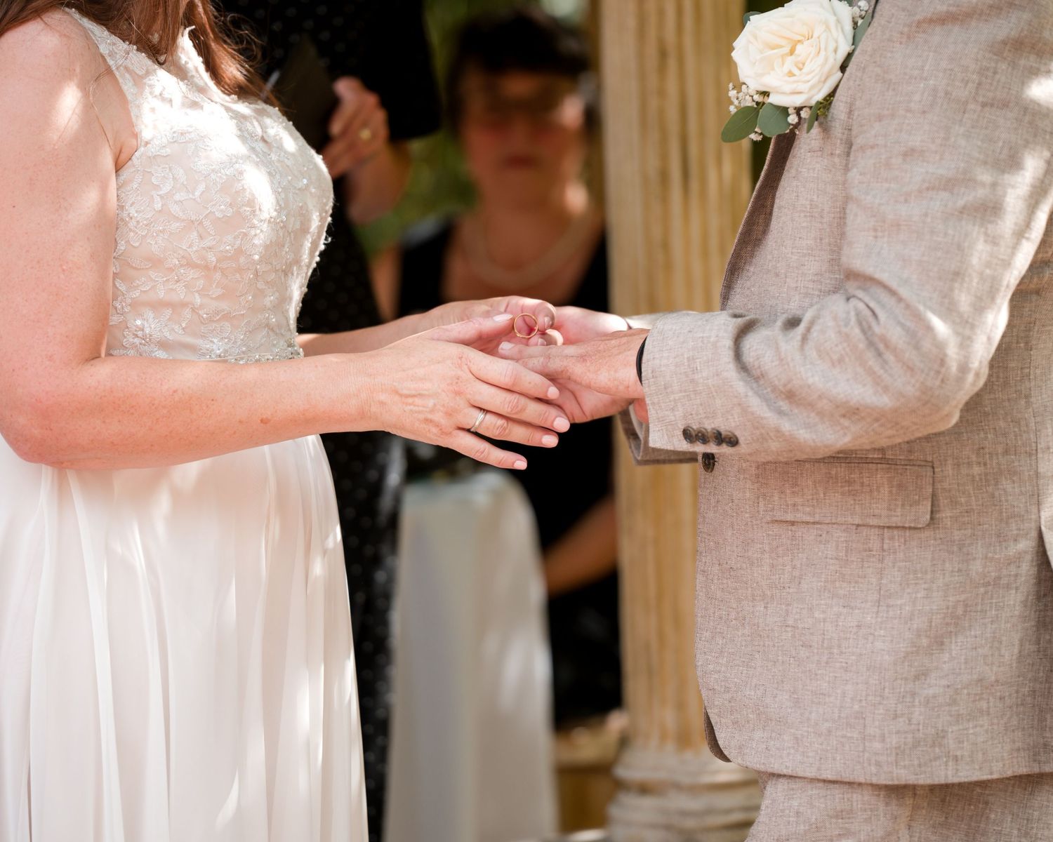 Bride and groom exchanging rings during outdoor wedding ceremony, groom wearing gray suit with white rose boutonniere.