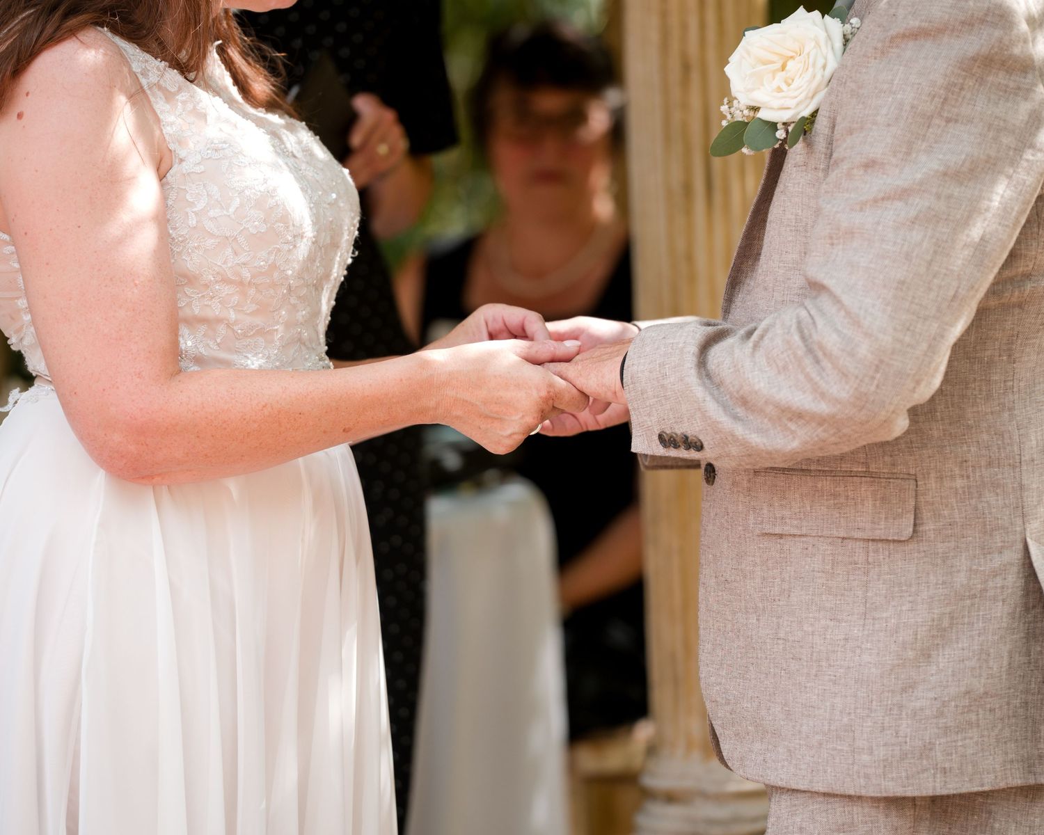 Bride and groom exchanging rings during wedding ceremony, bride in white lace gown, groom in beige suit with white rose boutonniere.